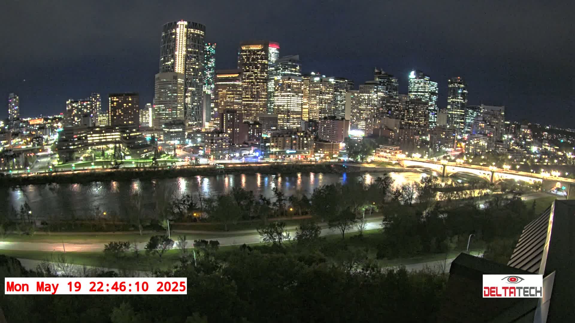 A brightly lit city skyline at night is situated across a calm river, surrounded by dark green trees under a clear sky.