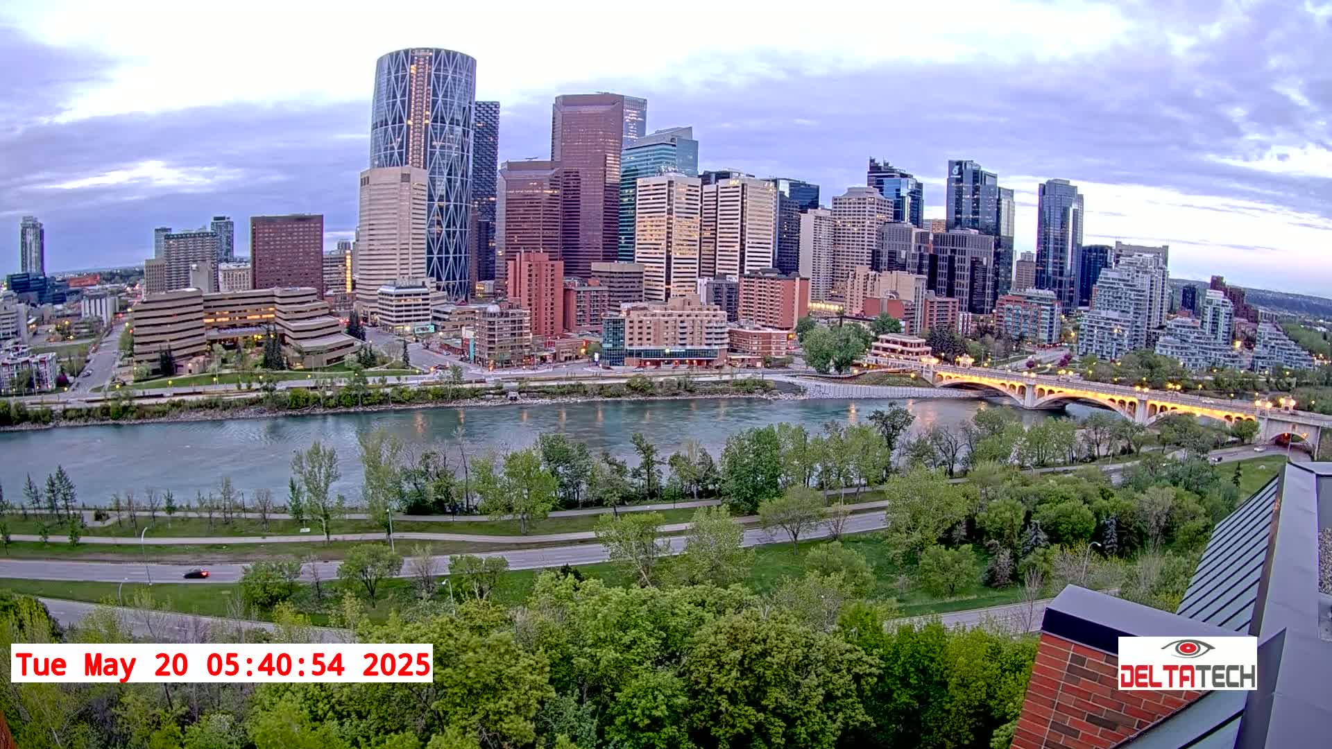 A city skyline with numerous skyscrapers and high-rise buildings sits across a river from a park with lush green trees under a partly cloudy sky.