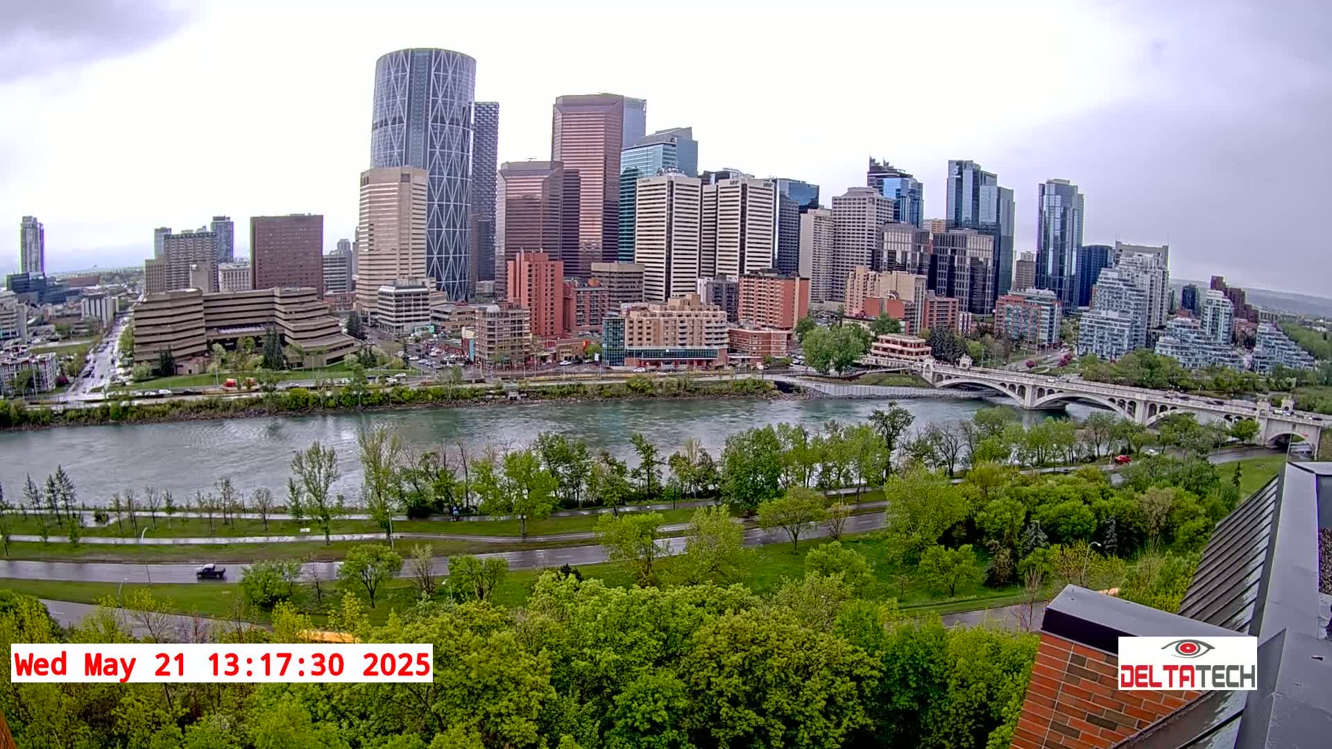 A city skyline with numerous high-rise buildings sits across a river from a lush green park under an overcast sky.