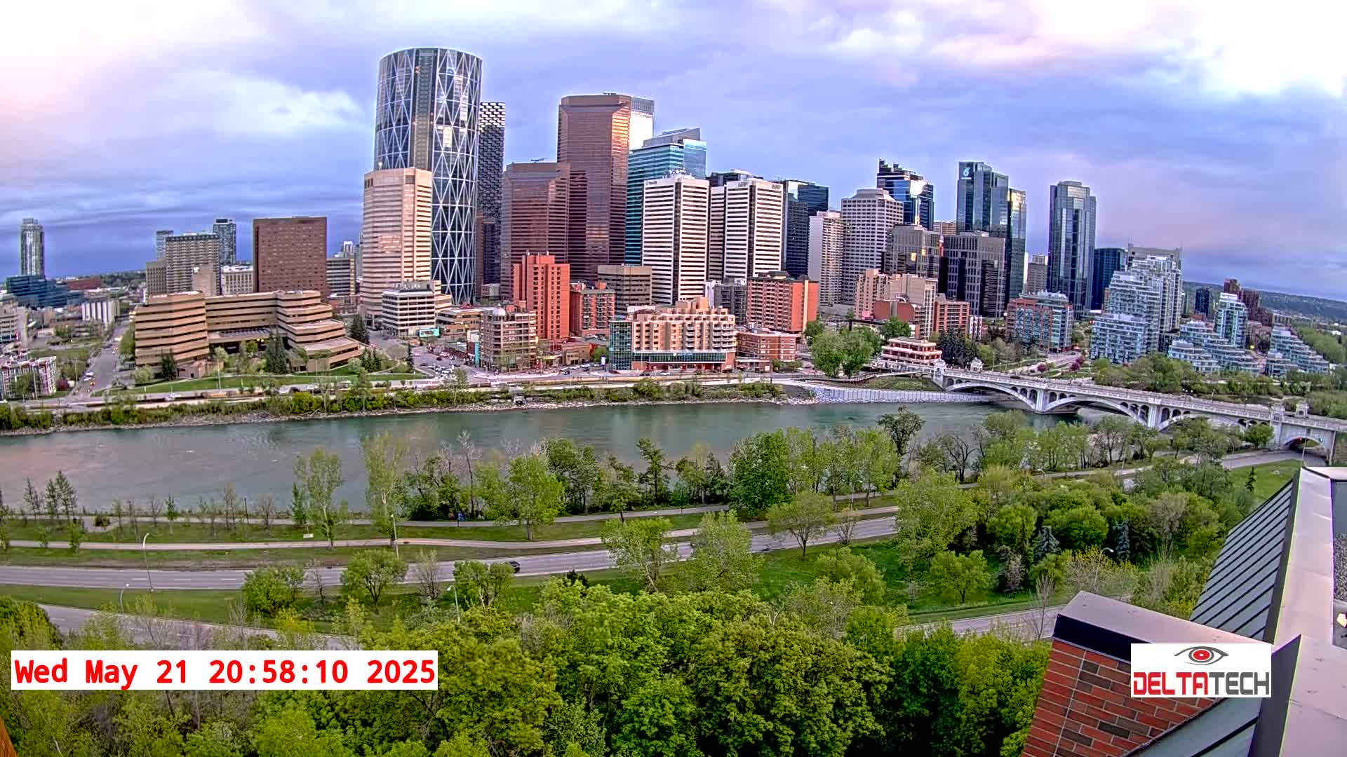 A city skyline with many high-rise buildings is seen across a river under a partly cloudy sky.