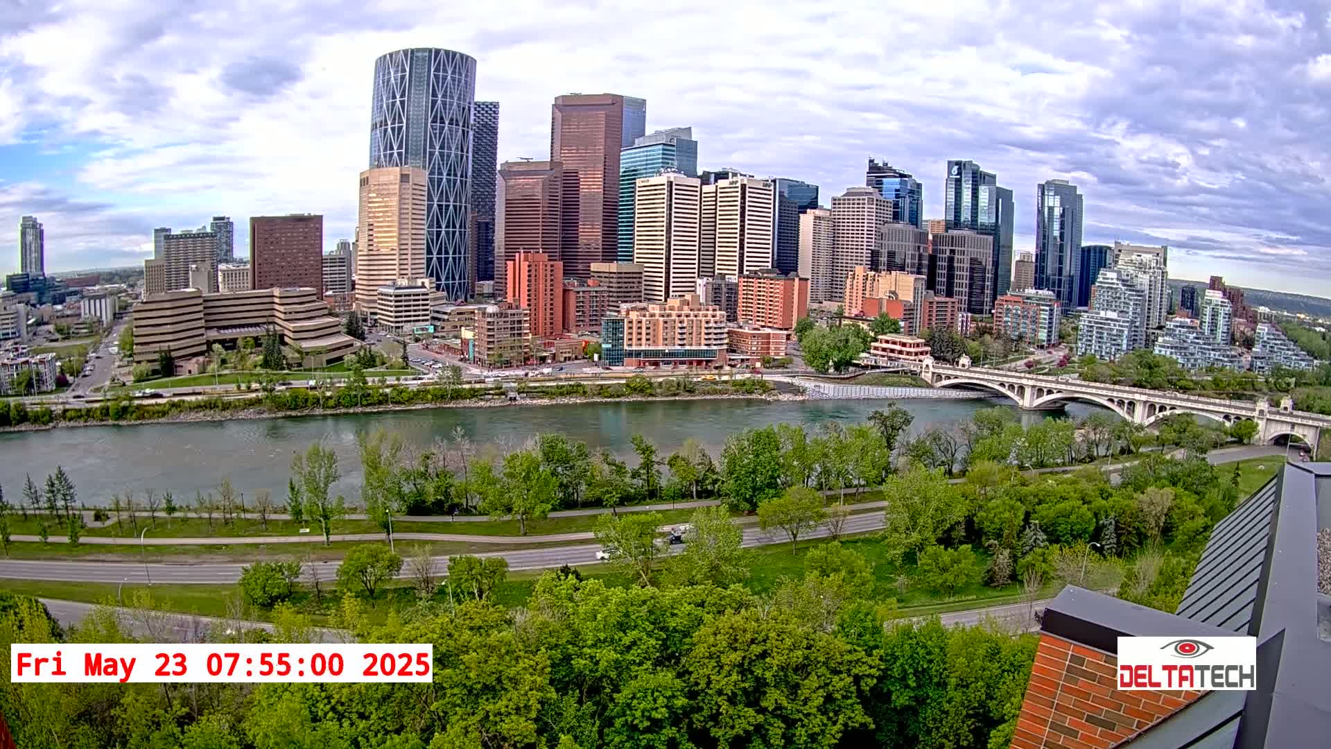 A city skyline with numerous skyscrapers is visible across a river, under partly cloudy skies, with lush green trees and a paved road in the foreground.
