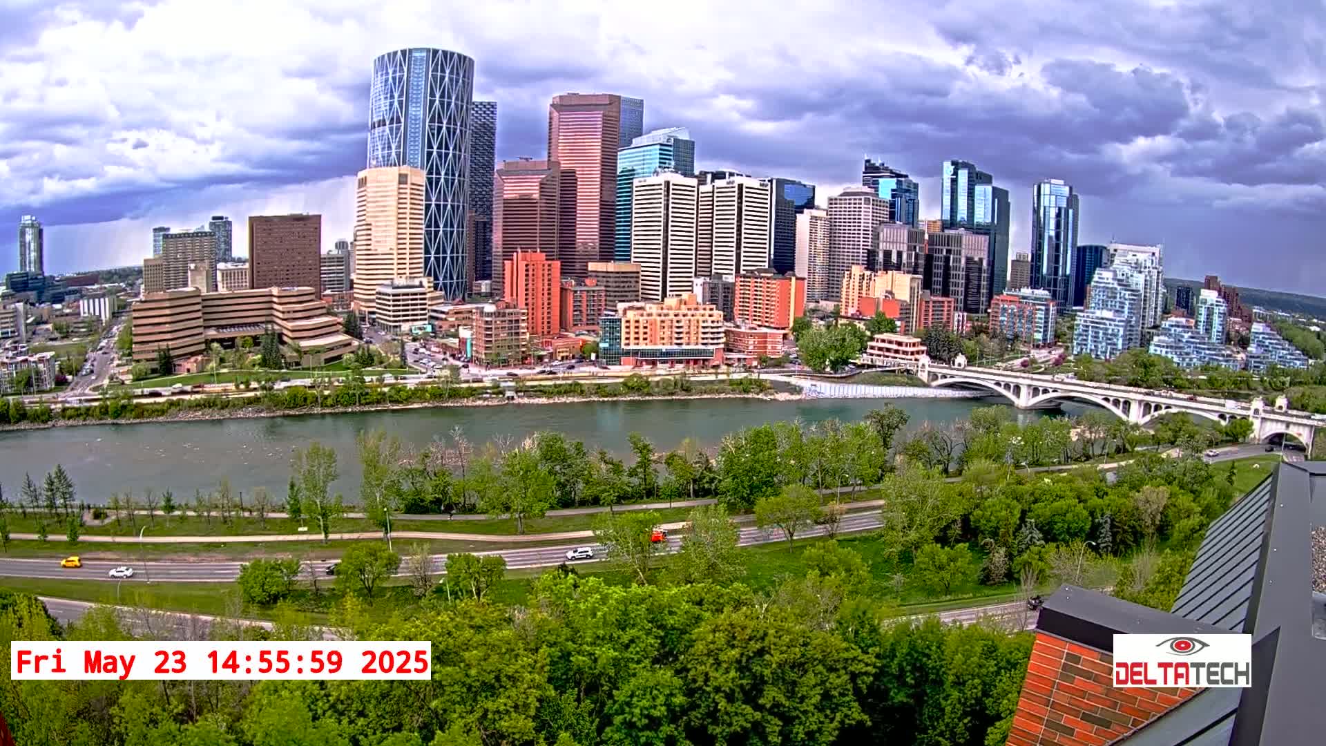 A city skyline with numerous skyscrapers is seen across a river under a partly cloudy sky, with green trees and a road leading to a bridge in the foreground.