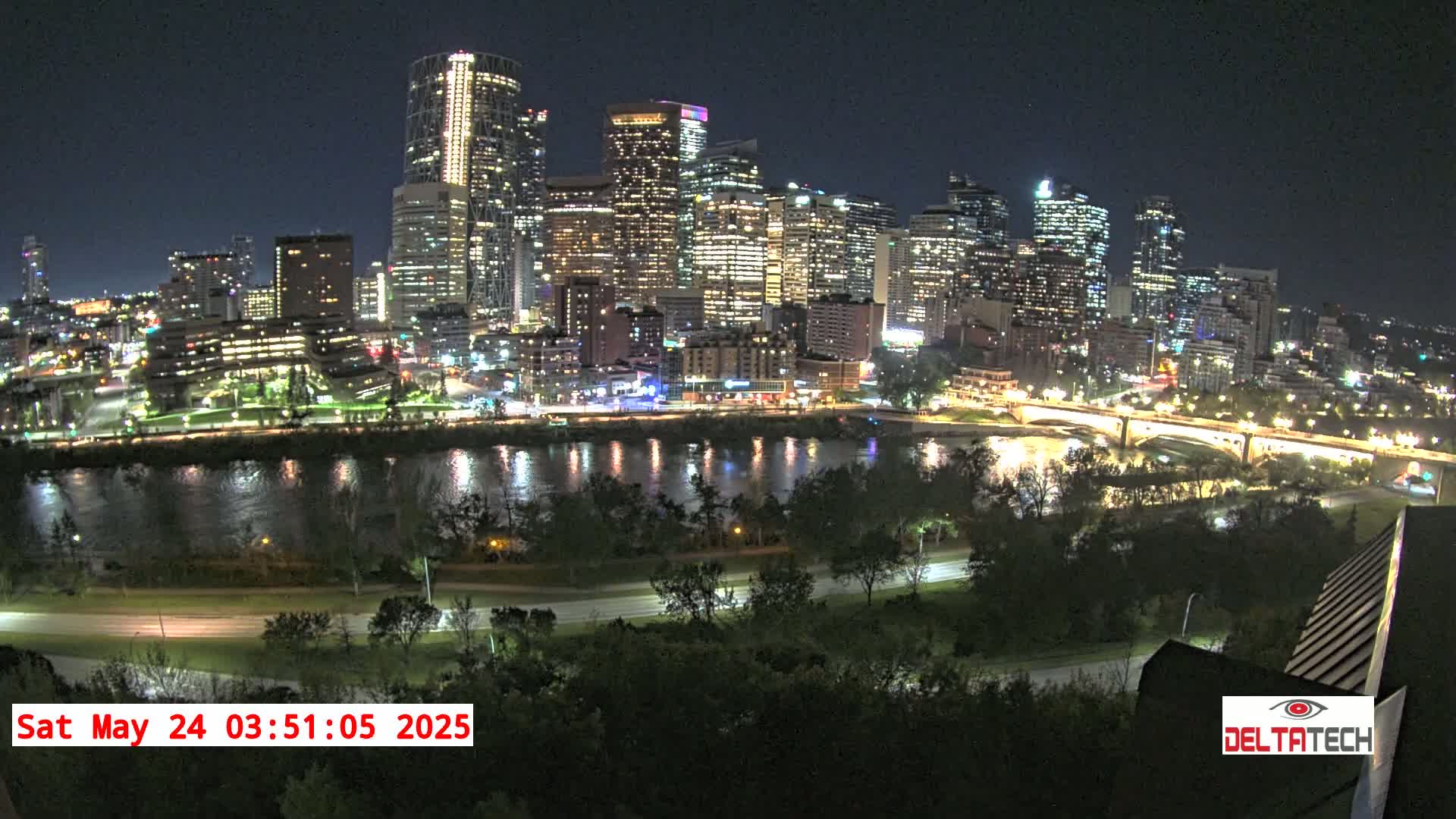 A brightly lit city skyline at night is seen across a dark river, under a clear sky.
