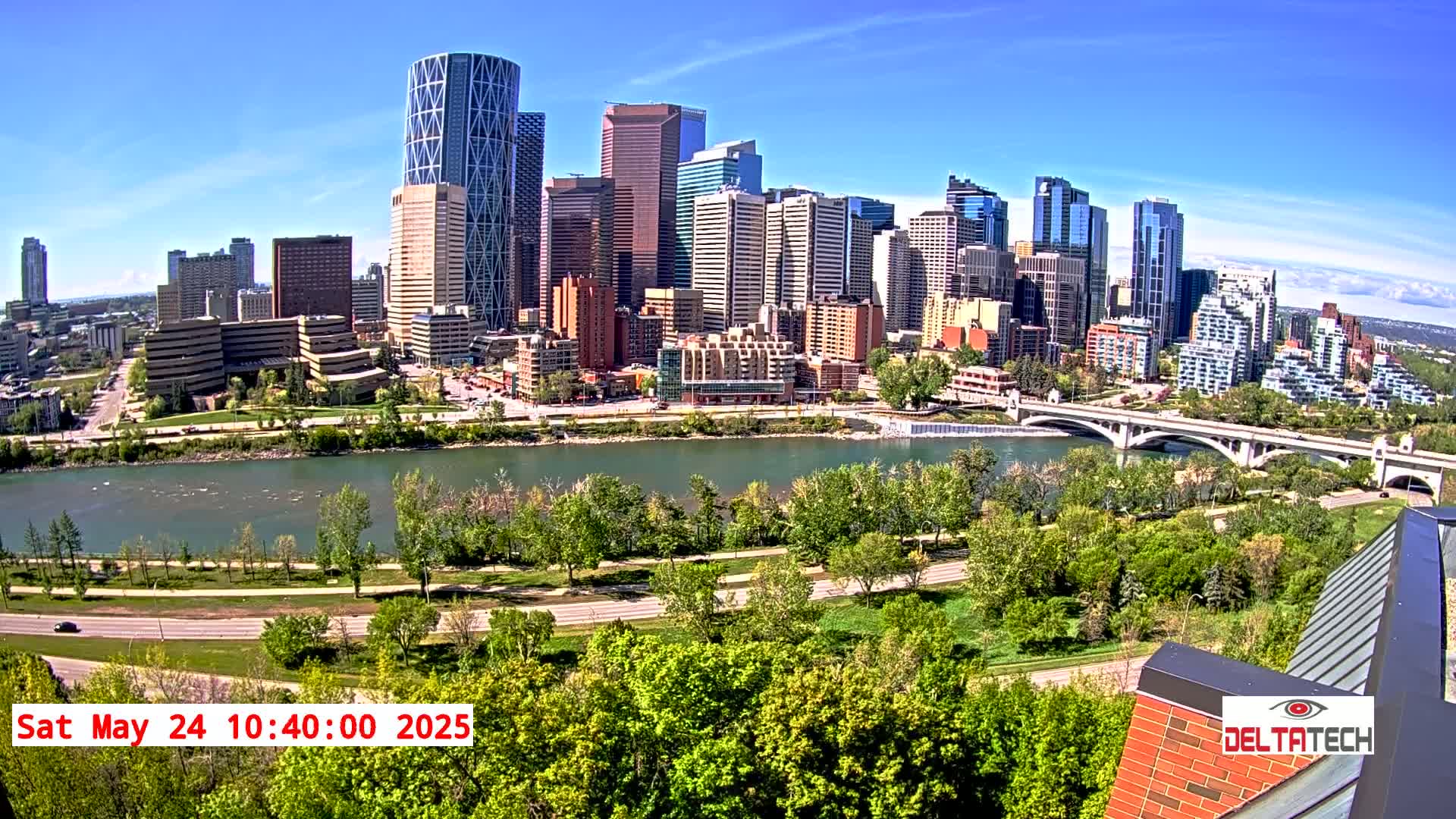 A sunny cityscape with a river and bridge is viewed from an elevated perspective, showing green trees along the riverbank and numerous high-rise buildings.