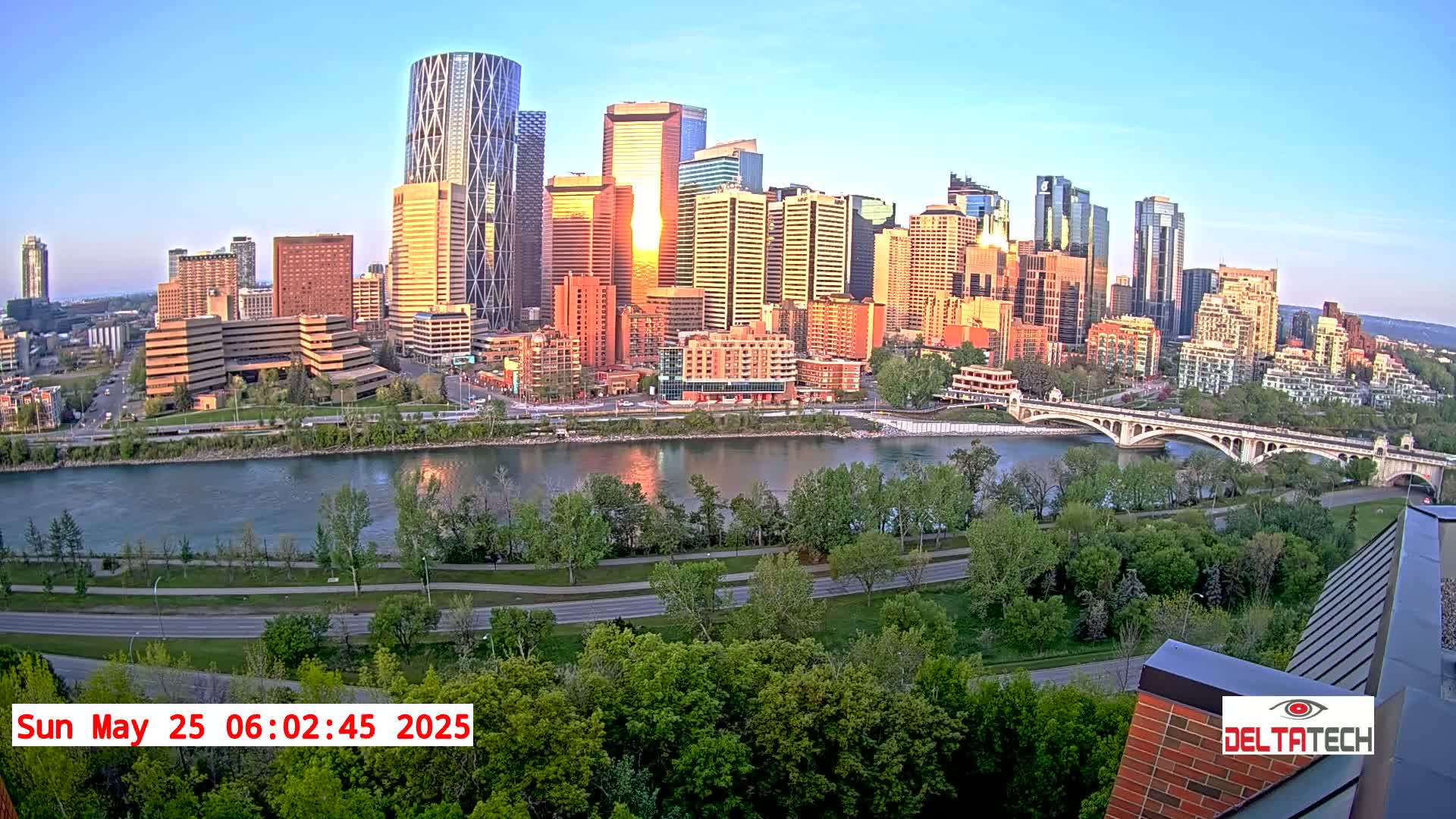 A city skyline with many tall buildings is seen across a river at sunrise, under a clear sky.