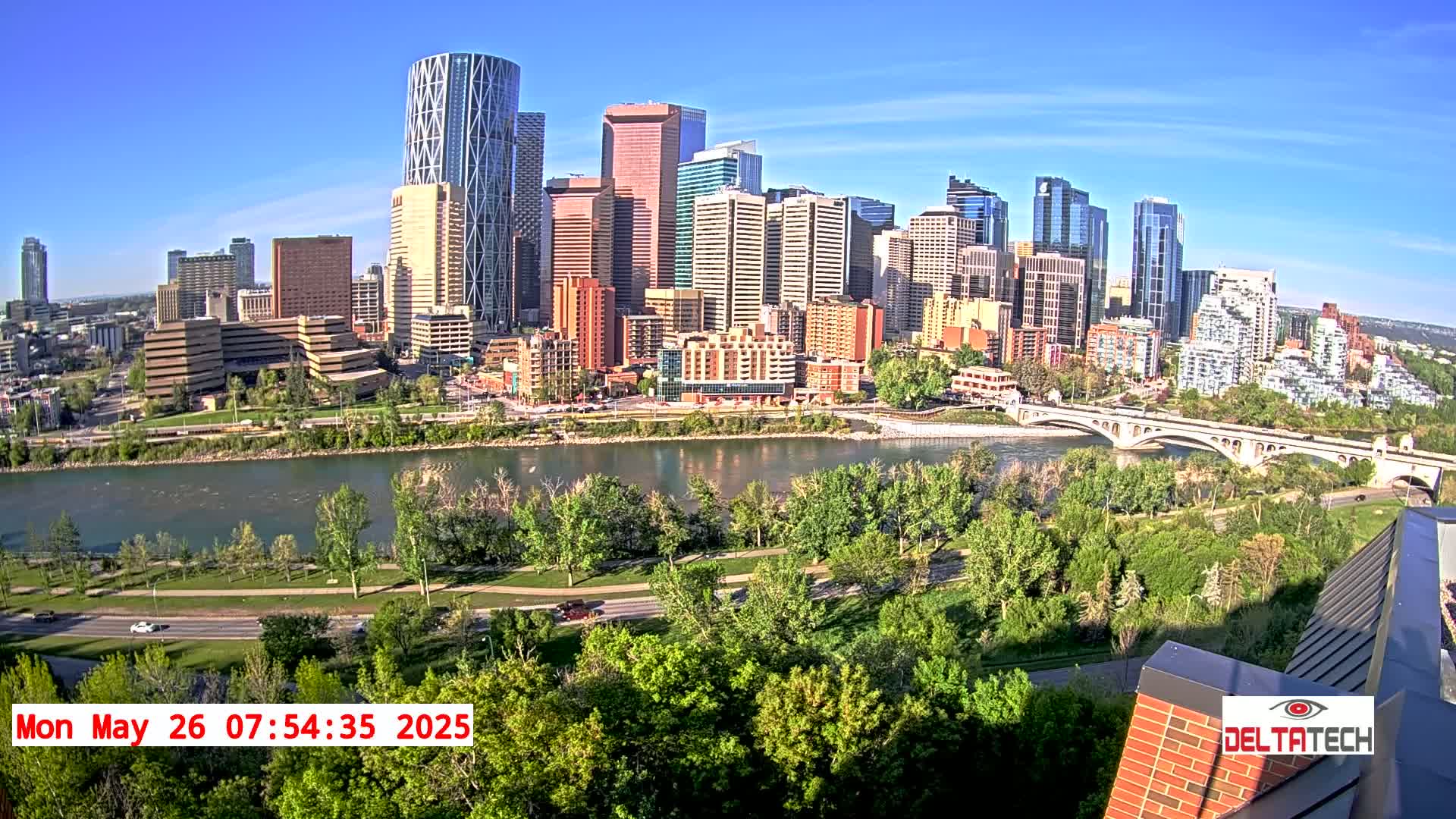A city skyline with many tall buildings is seen across a river on a sunny day, with lush green trees lining the riverbank and a bridge in the mid-ground.