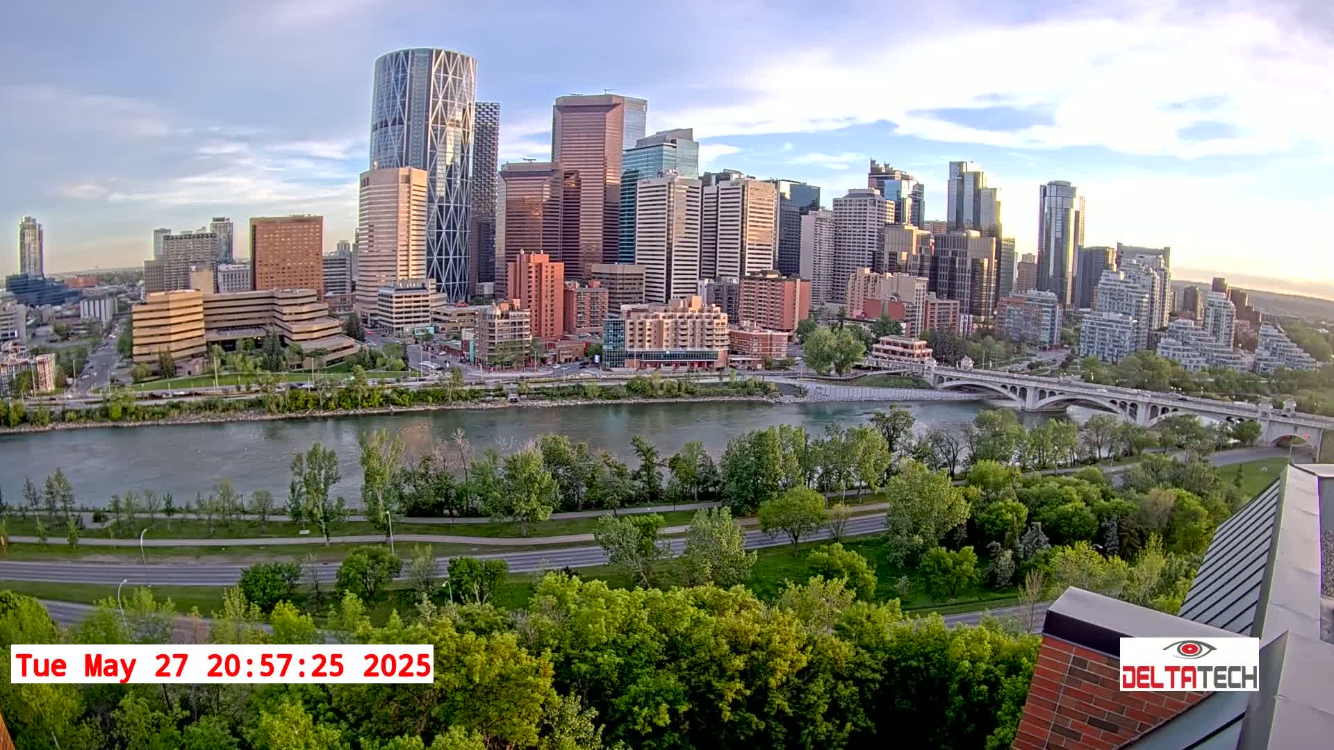 A city skyline with many tall buildings sits on the far side of a wide river, under a partly cloudy sky.