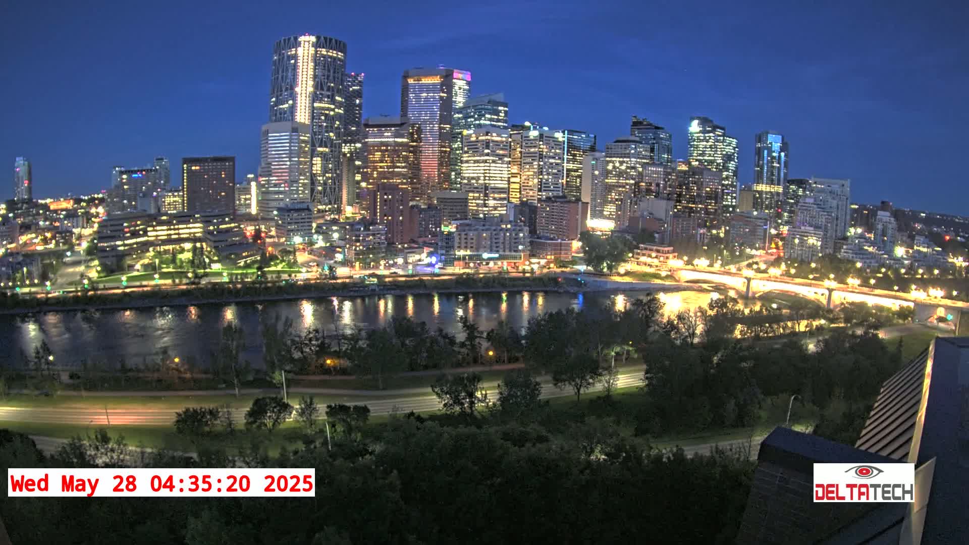 A brightly lit city skyline at night is situated across a calm river, bordered by green trees and roads.