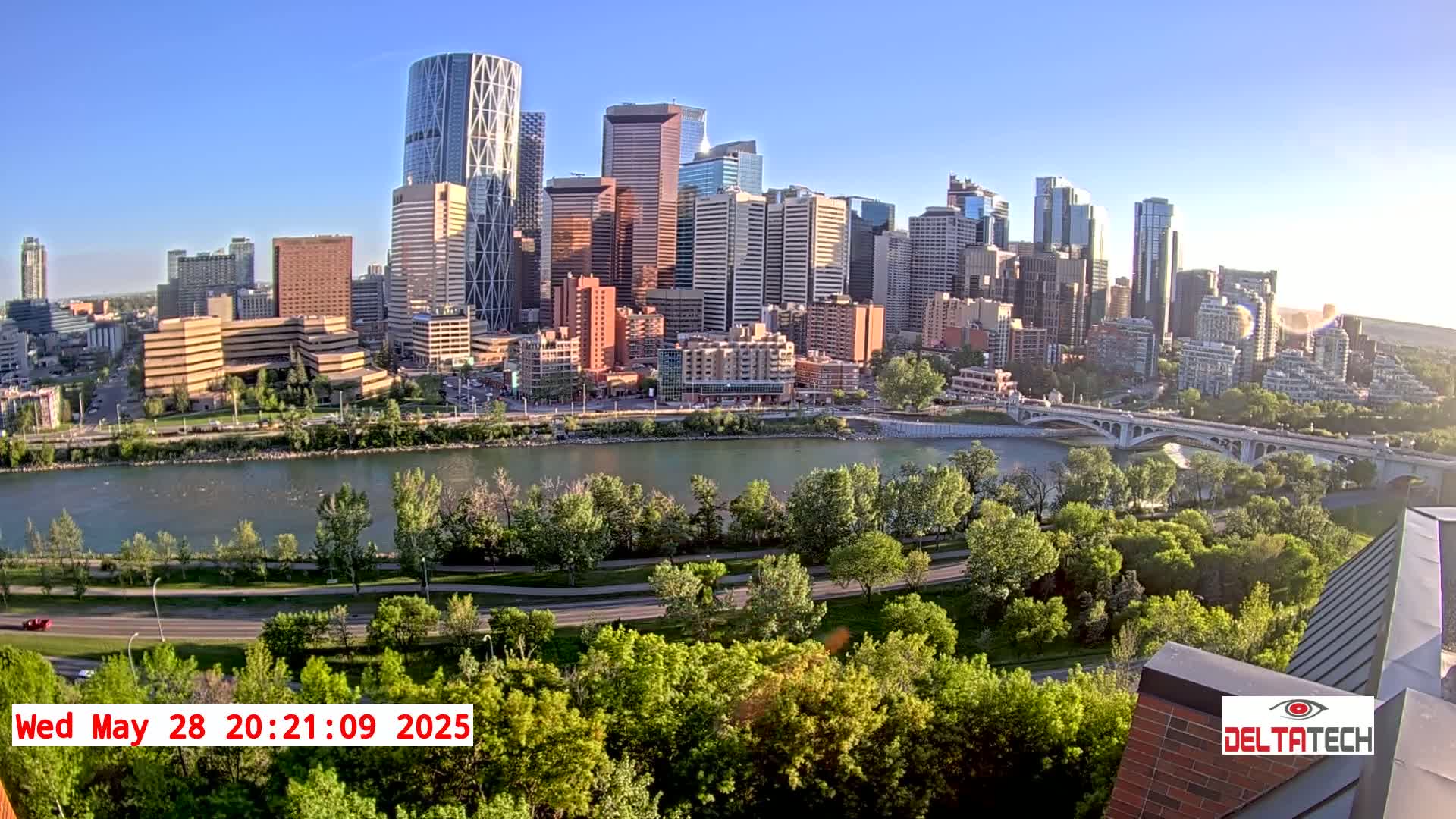 A cityscape with numerous skyscrapers is viewed from across a river, under a clear, sunny sky, with lush green trees lining the riverbanks and roads.