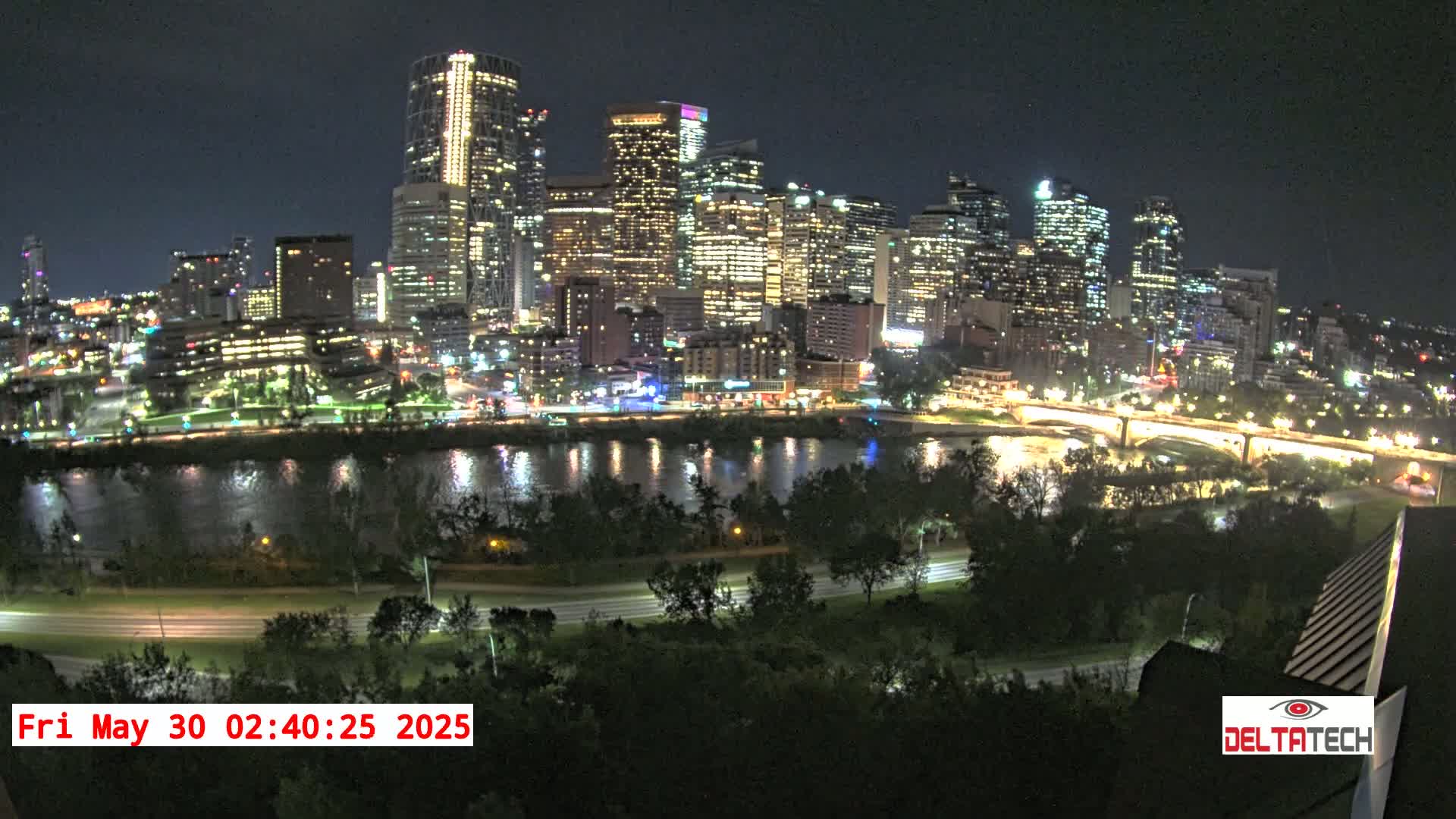 A brightly lit city skyline at night is seen across a dark river, with trees and roads in the foreground under a clear sky.