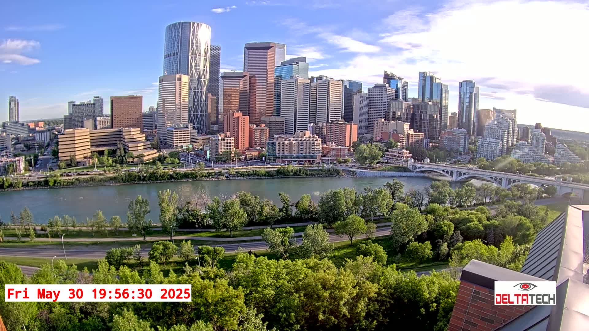 A city skyline with many tall buildings is situated across a river from a park with lush green trees, under a mostly sunny sky.