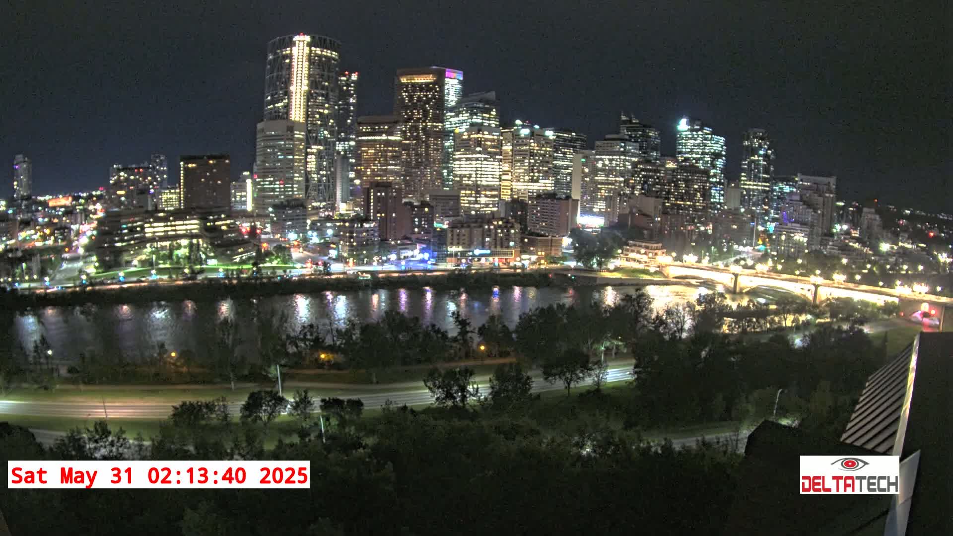 A brightly lit city skyline at night is seen across a river from a high vantage point under a clear, dark sky.