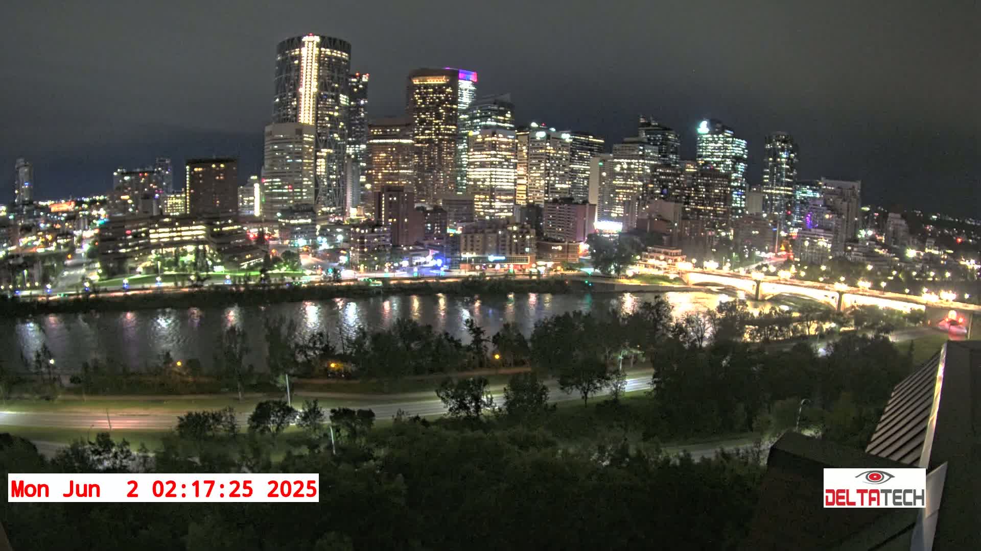 A brightly lit city skyline at night is seen across a dark river, with lush green trees and roads in the foreground under a dark sky.
