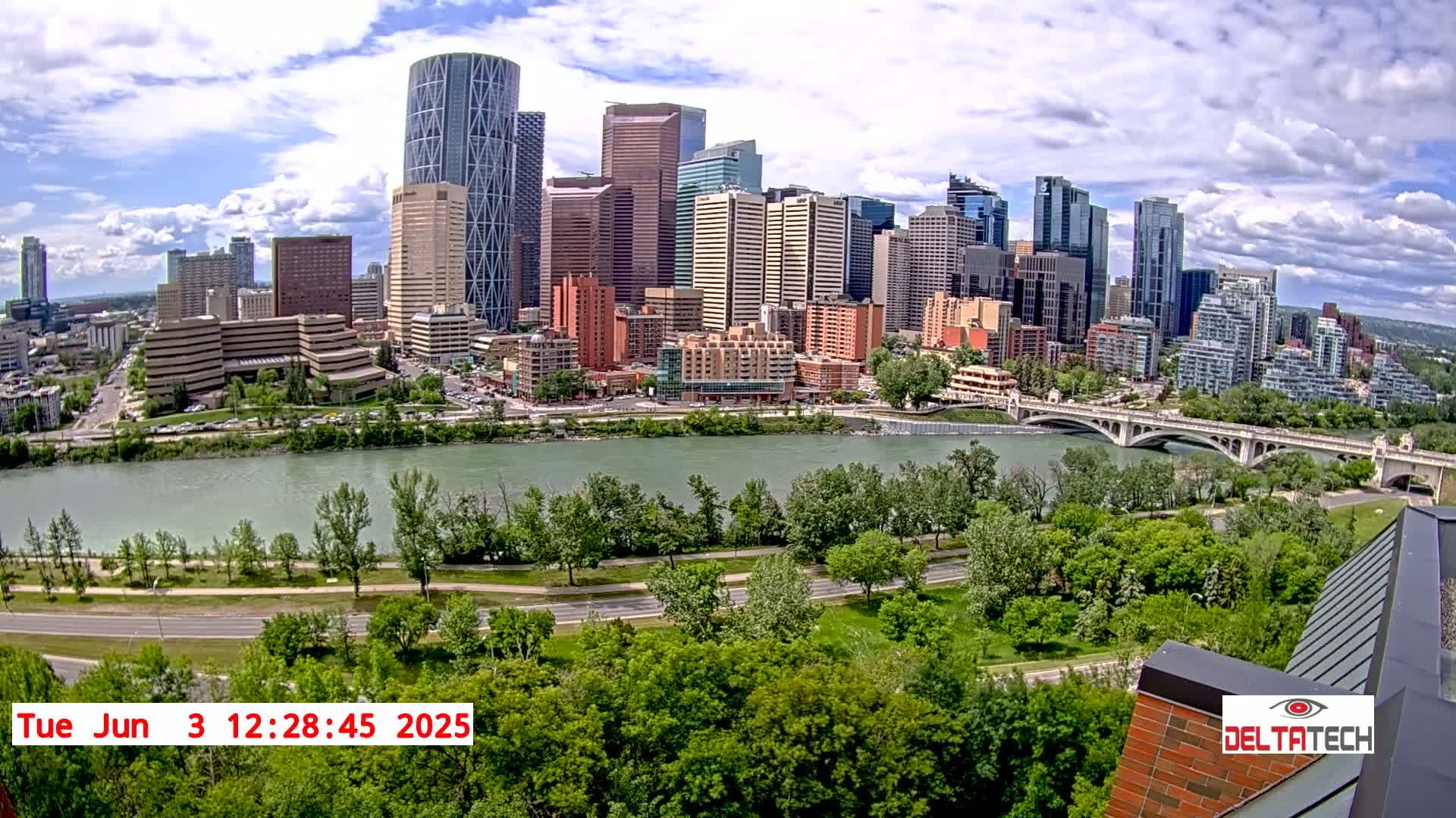 A city skyline with numerous skyscrapers is viewed from across a river on a partly cloudy day, with lush green trees lining the riverbank and a bridge spanning the waterway.