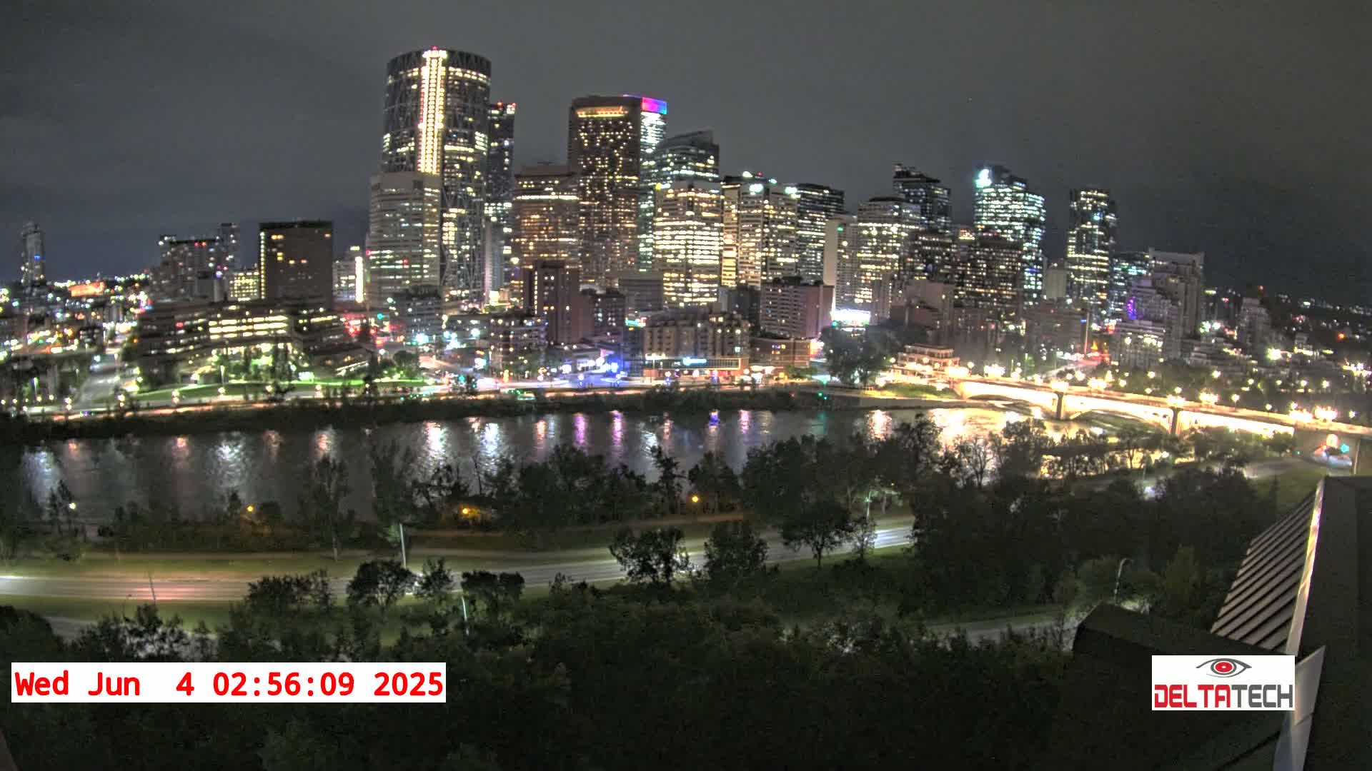 A nighttime view of a city skyline illuminated by lights, across a dark river, with trees and roads in the foreground.