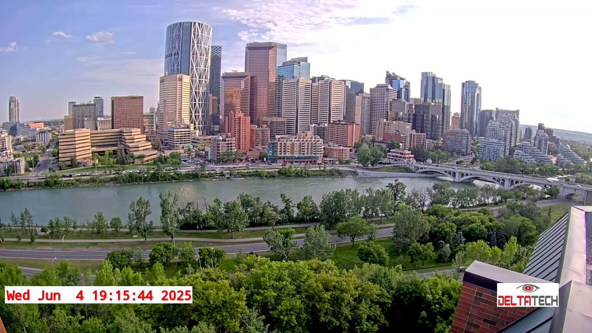 A cityscape with numerous skyscrapers and high-rise buildings is situated across a river from a park-like area with lush green trees and a roadway, under partly cloudy skies.