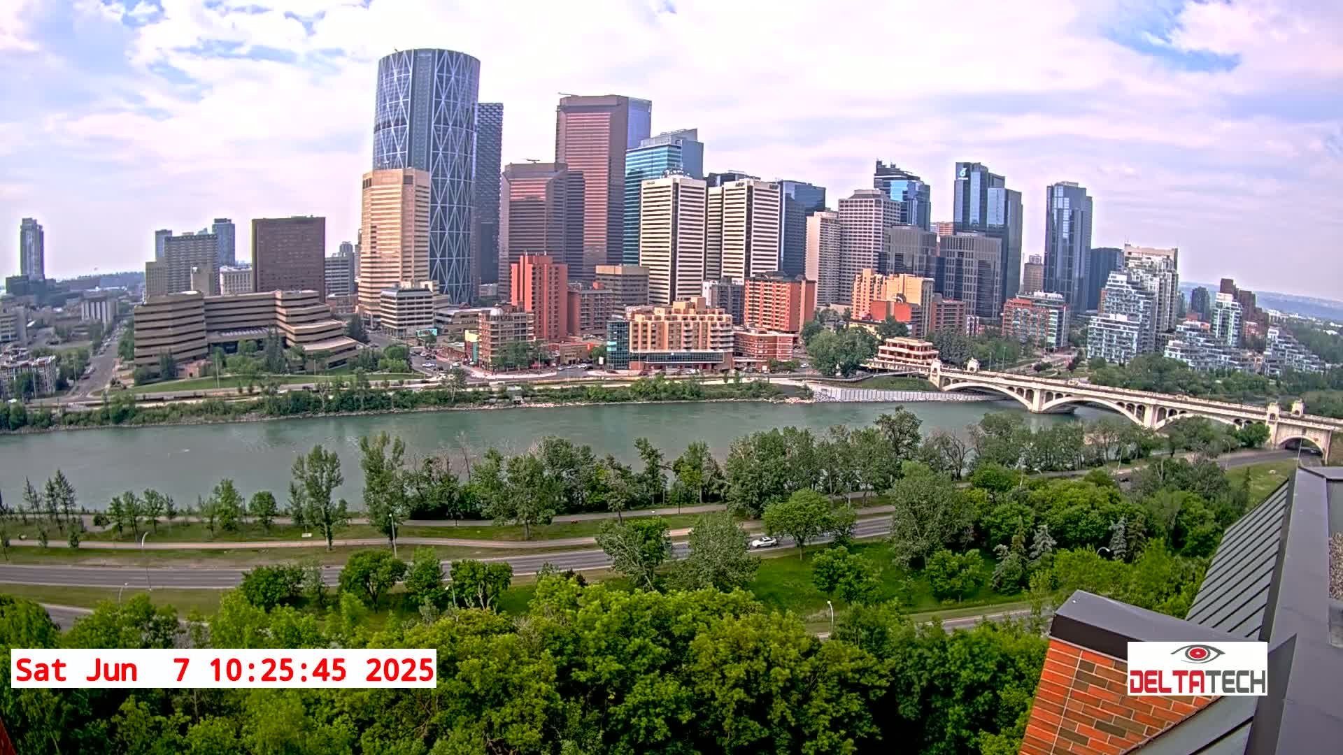 A city skyline with many tall buildings sits across a river from a park with lush green trees under a partly cloudy sky.