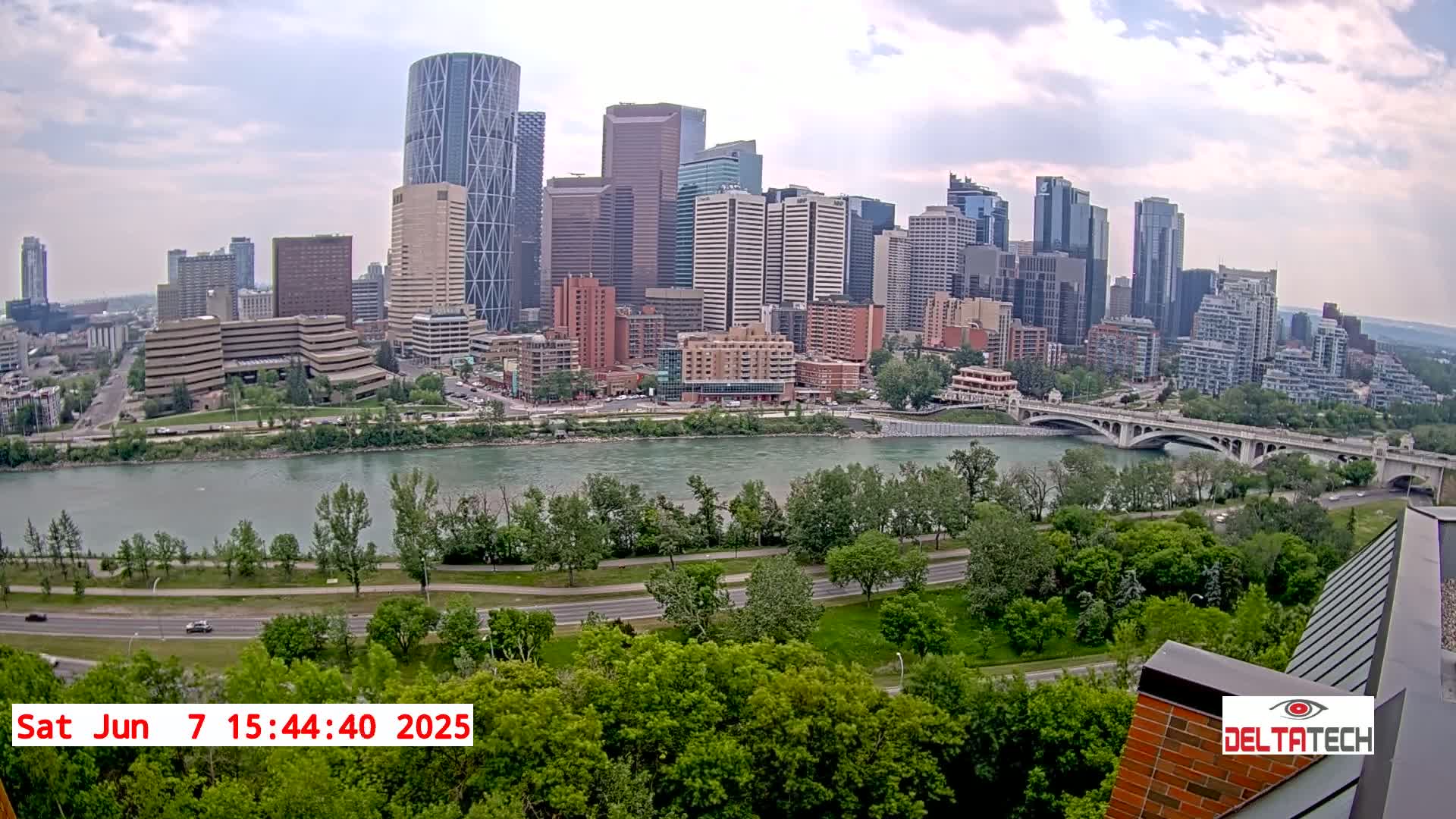 A city skyline with numerous skyscrapers is visible across a wide river, under a partly cloudy sky, with lush green trees and a roadway in the foreground.