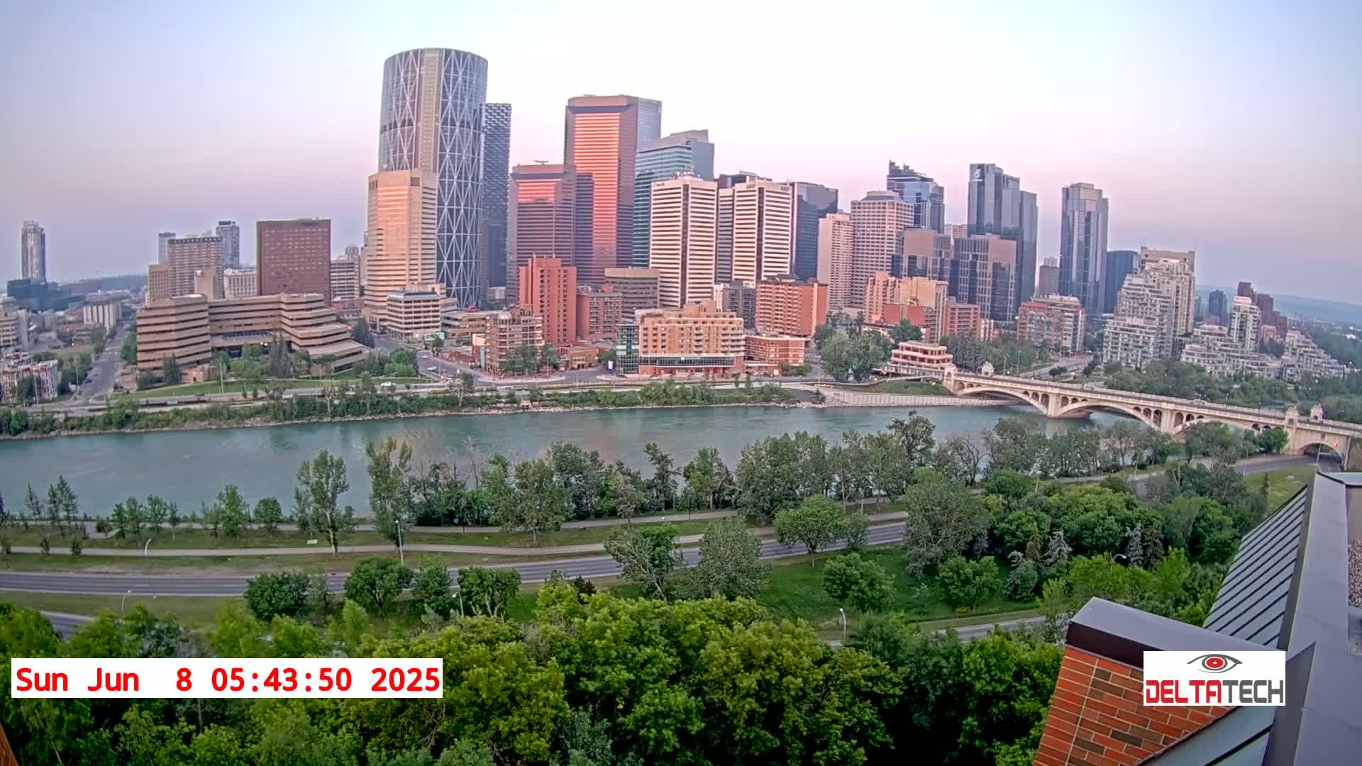 A city skyline with numerous high-rise buildings sits across a river from a bridge under a light-grey sky.