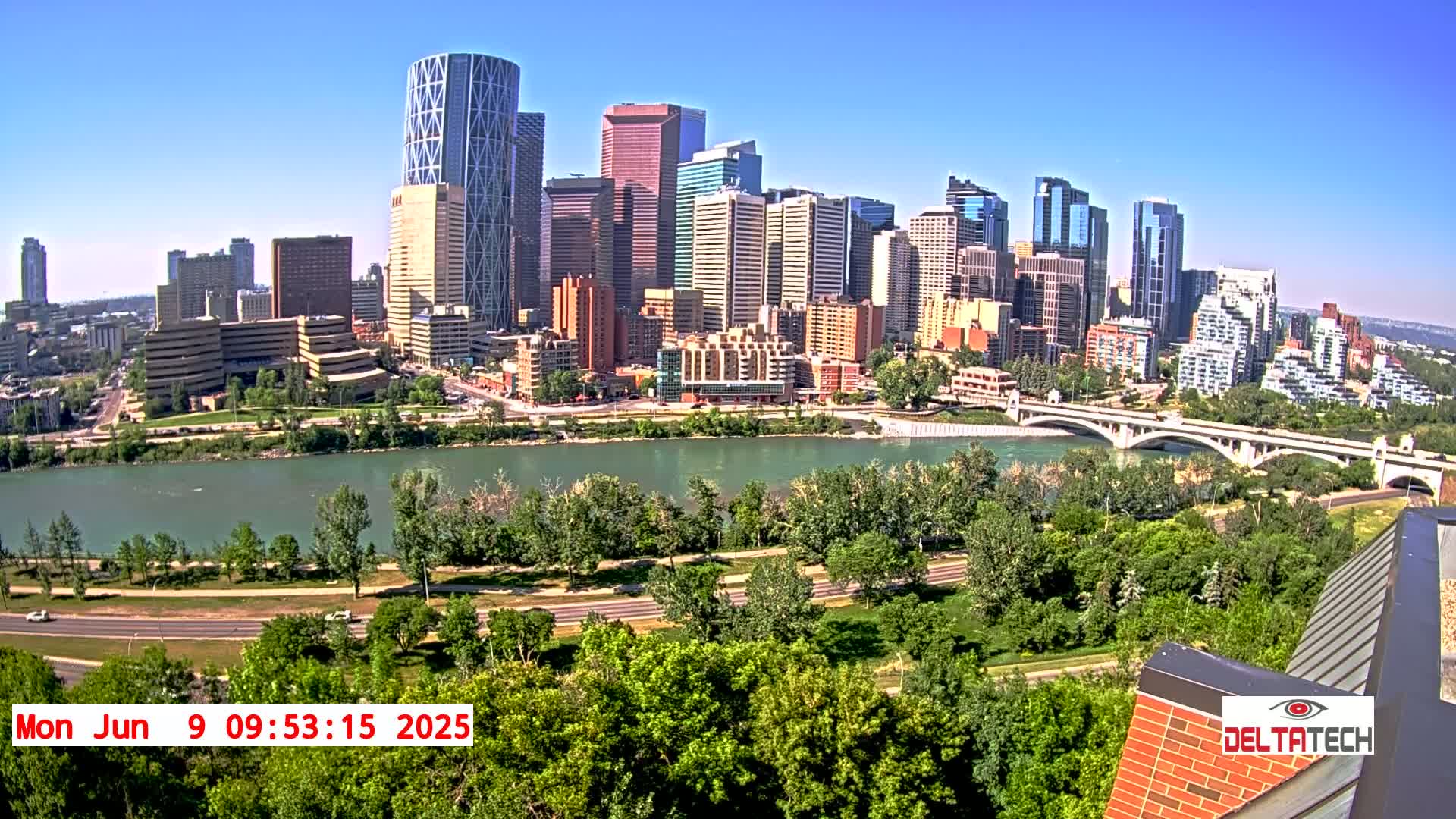 A sunny cityscape with numerous skyscrapers and high-rise buildings sits across a wide river from a lush green park, a roadway, and a bridge.