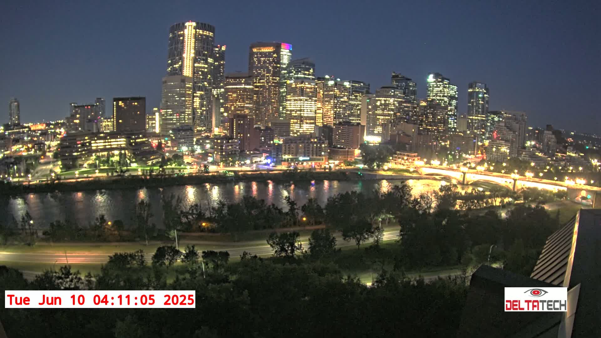 A brightly lit city skyline at night is situated across a river from a dark, tree-lined park.