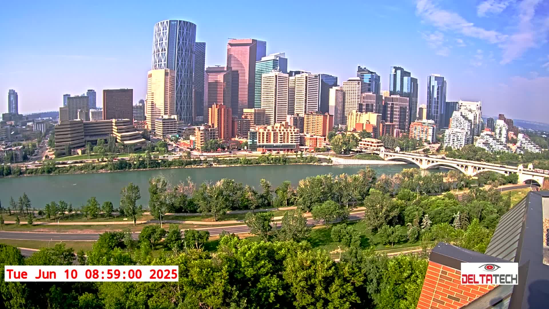 A city skyline with many tall buildings is situated across a river from lush green parkland on a sunny day.