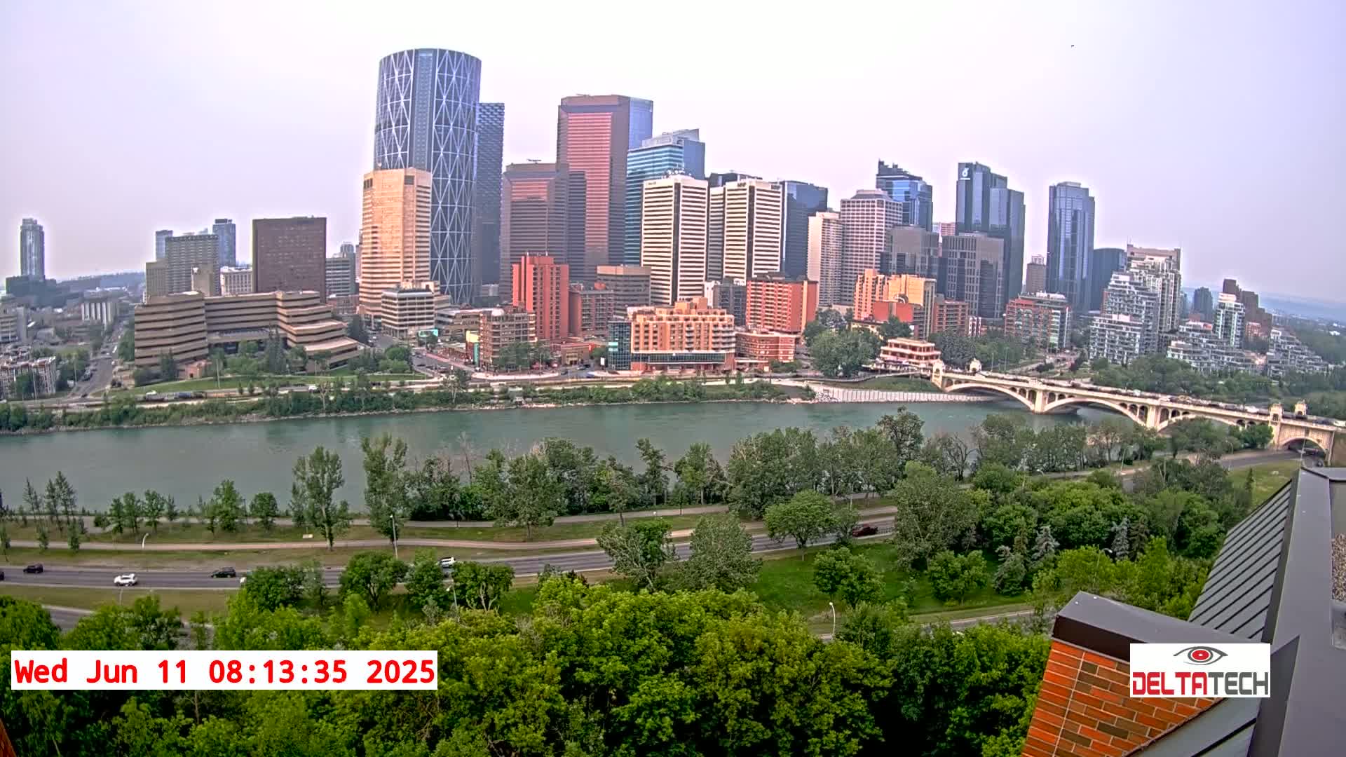 A hazy cityscape with numerous skyscrapers sits across a wide river from a green park and roadway, with a bridge connecting the city to the other side.