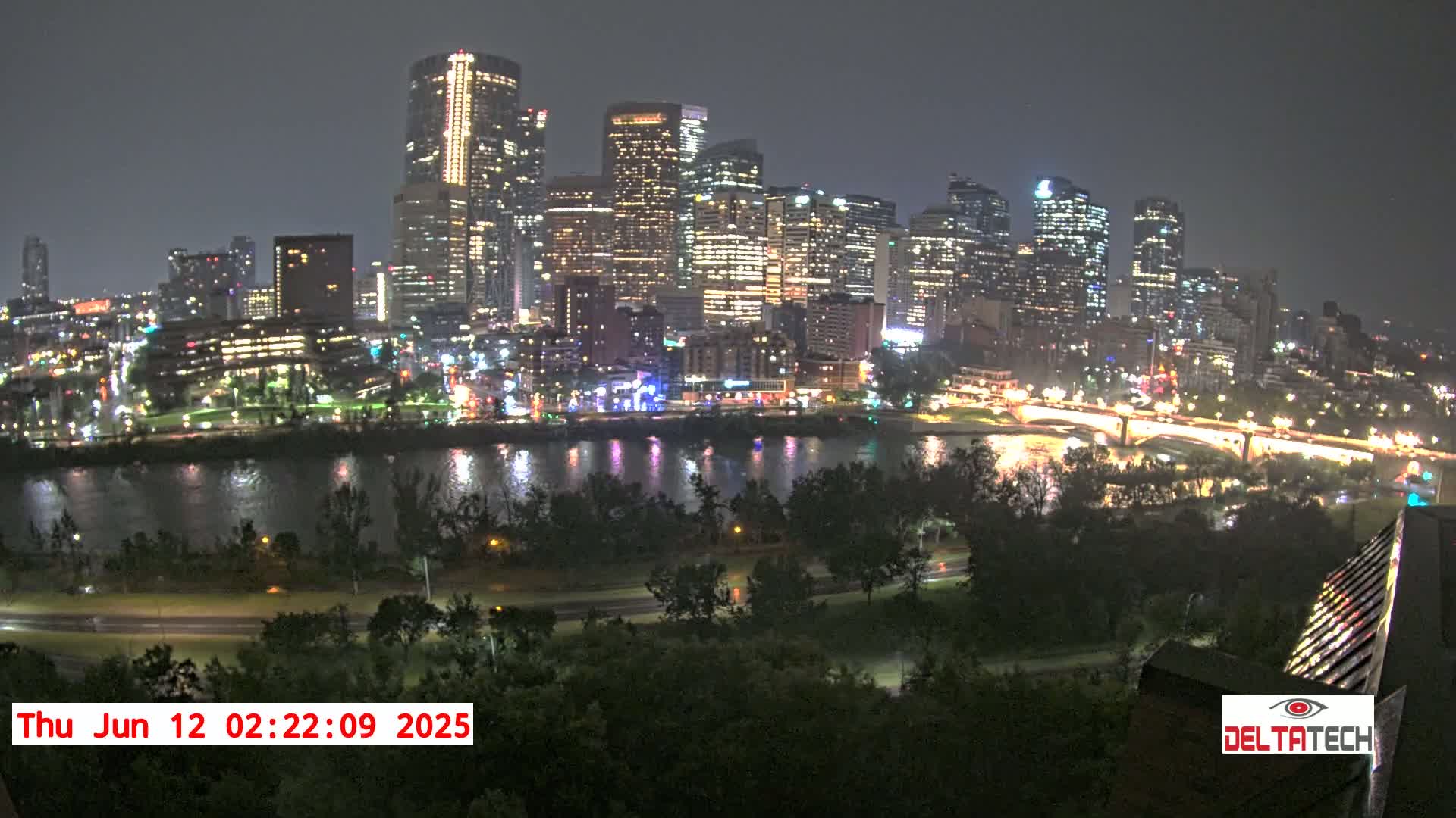 A brightly lit city skyline at night is seen across a dark river from a high vantage point under a dark, possibly overcast sky.