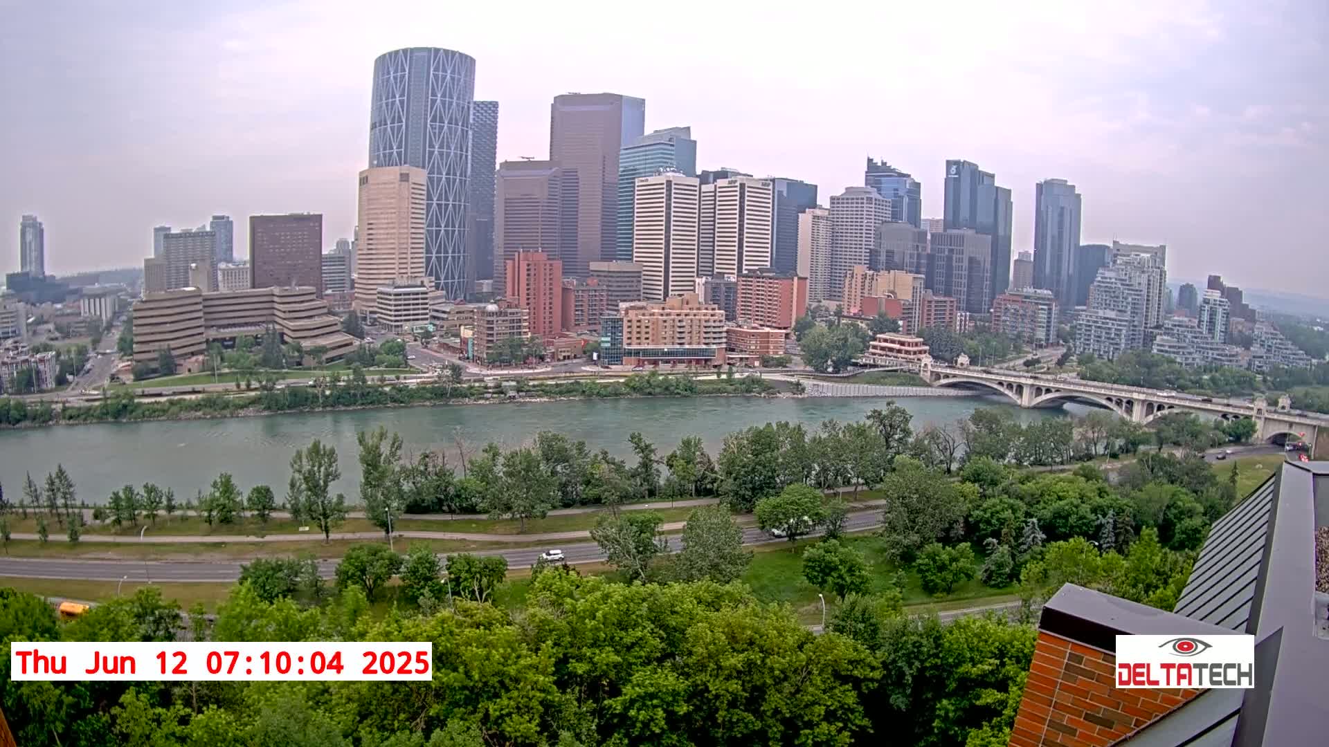 A cityscape with numerous skyscrapers sits across a wide river from a lush green park, under an overcast sky.