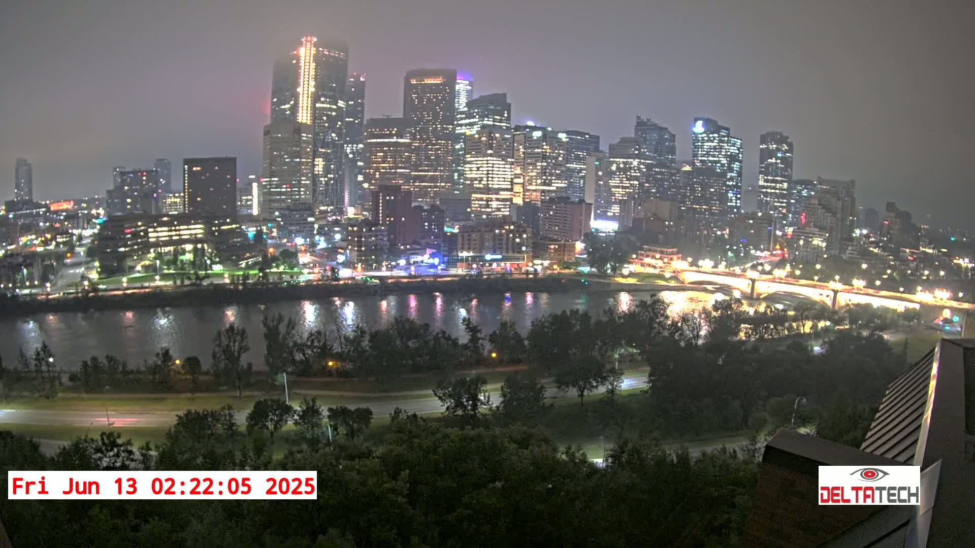 A nighttime cityscape with many illuminated buildings is seen across a river, under hazy or foggy conditions.