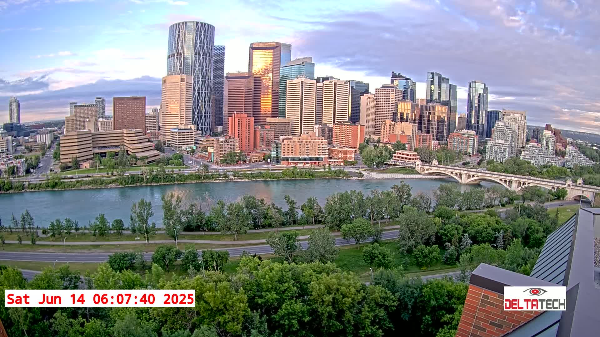 A city skyline with numerous high-rise buildings is situated across a river under a partly cloudy, sunrise sky.