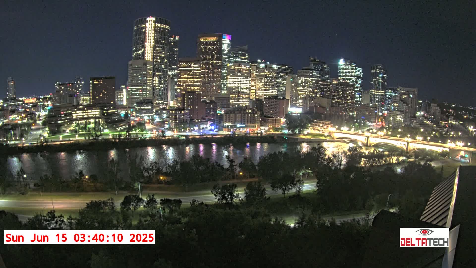 A nighttime view of a brightly lit city skyline across a calm river, with trees and roads in the foreground under a clear, dark sky.