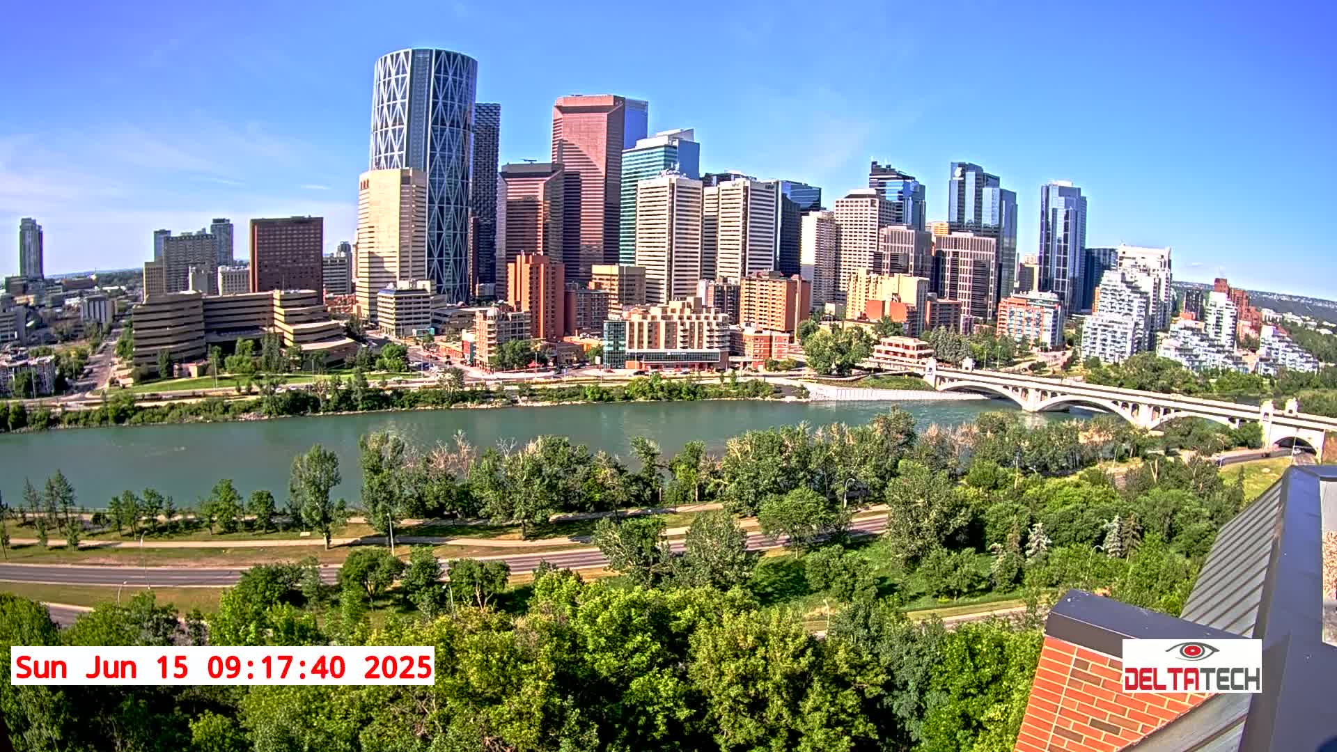 A city skyline with many tall buildings is visible across a wide river on a sunny day, with lush green trees lining the riverbanks and a bridge spanning the water.