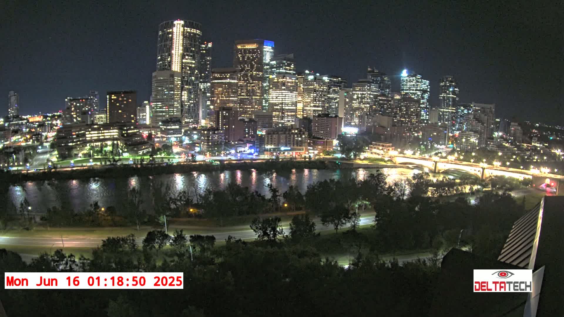 A brightly lit city skyline at night is seen across a dark river, with a road and trees in the foreground under a clear night sky.