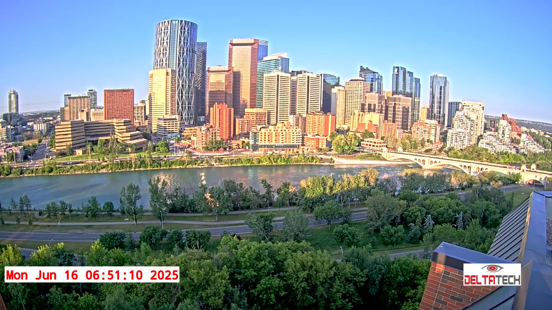 A city skyline with numerous high-rise buildings sits across a river from a lush green park under a clear, sunny sky.