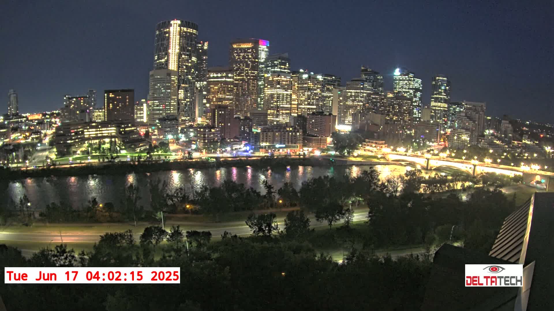 A brightly lit city skyline at night is seen across a dark river, under a clear sky.