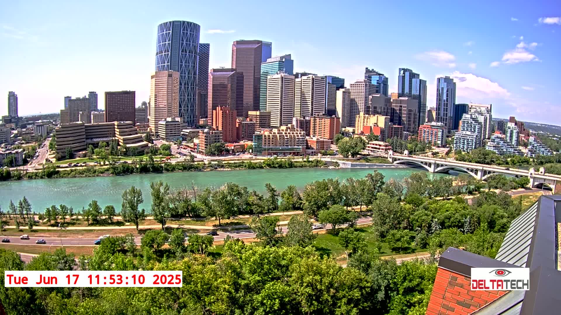 A city skyline with numerous skyscrapers is seen across a wide river on a sunny day, with lush green trees lining the riverbanks and a bridge spanning the waterway.
