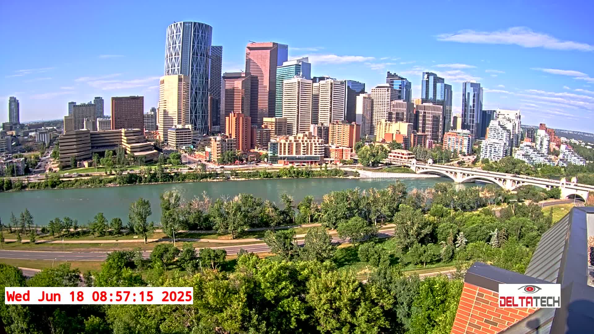A city skyline with numerous skyscrapers is visible across a river, under sunny skies, with lush green trees and a bridge in the foreground.