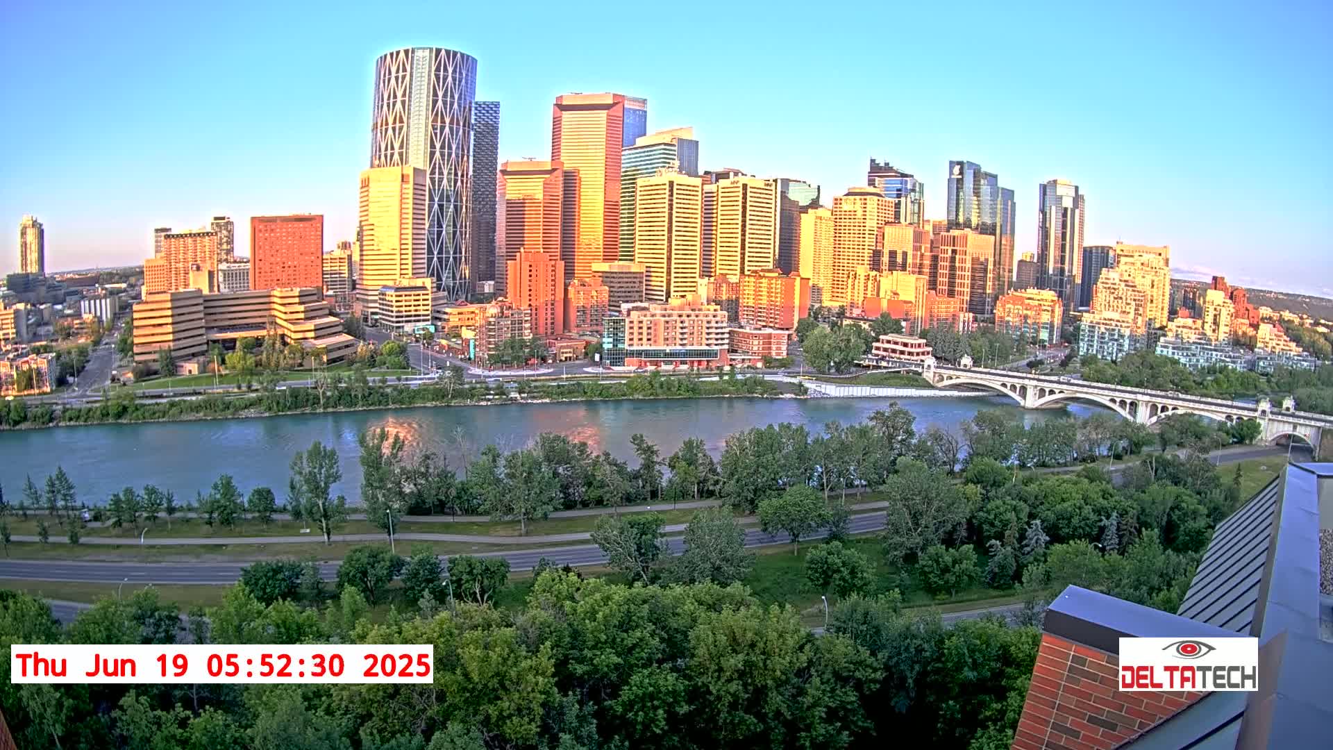 A cityscape at sunrise, showing a river with a bridge and a cityscape of tall buildings bathed in warm sunlight, surrounded by lush green trees and a roadway.