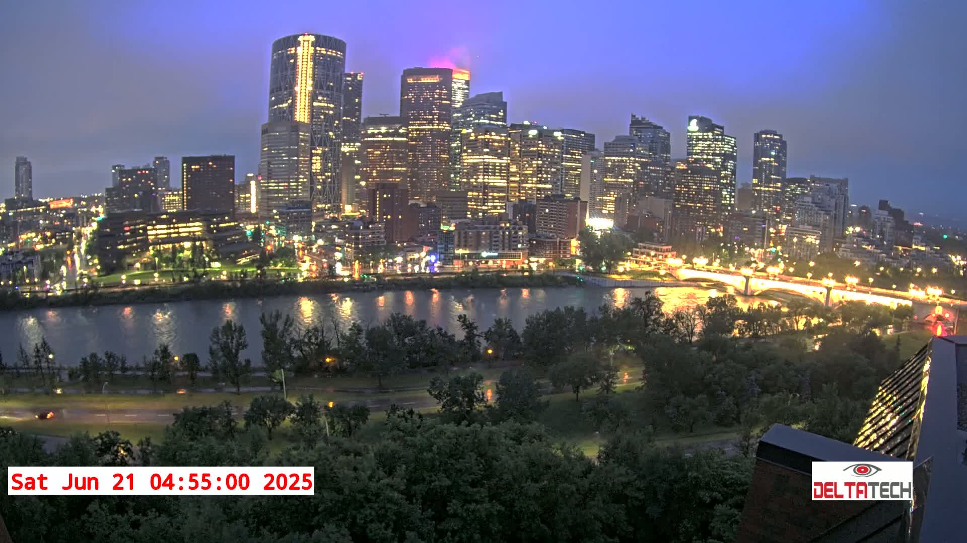 A brightly lit city skyline at twilight is seen across a calm river, with lush green trees and roads in the foreground.