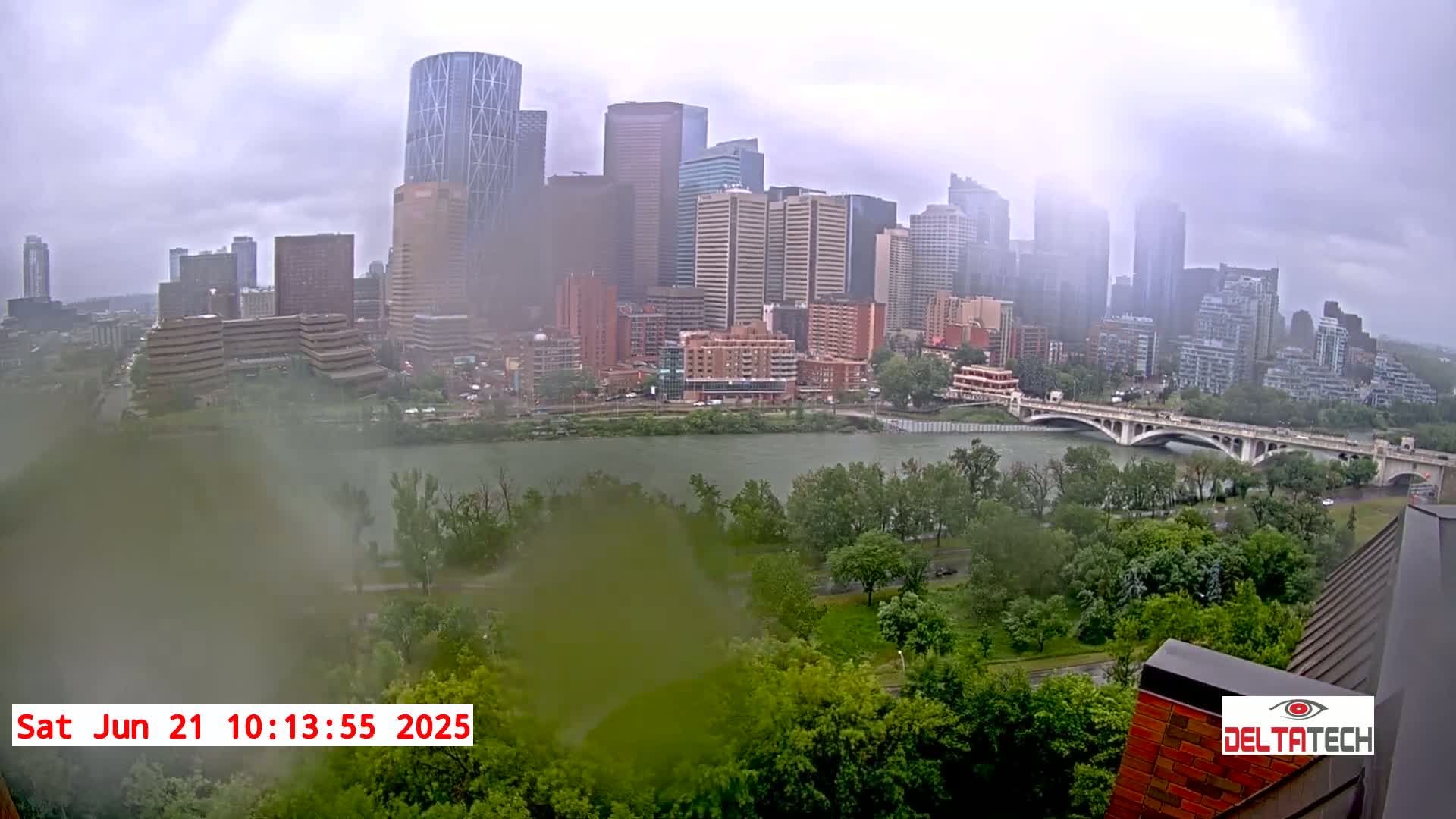 A city skyline with numerous high-rise buildings is viewed from across a river on a cloudy, rainy day.