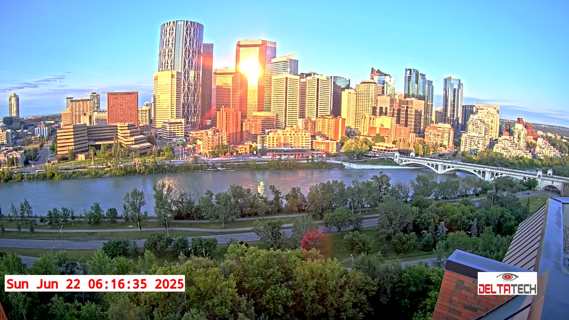 A cityscape with numerous skyscrapers and high-rise buildings is situated along a river, with a bridge crossing the river, under a clear, sunrise sky.