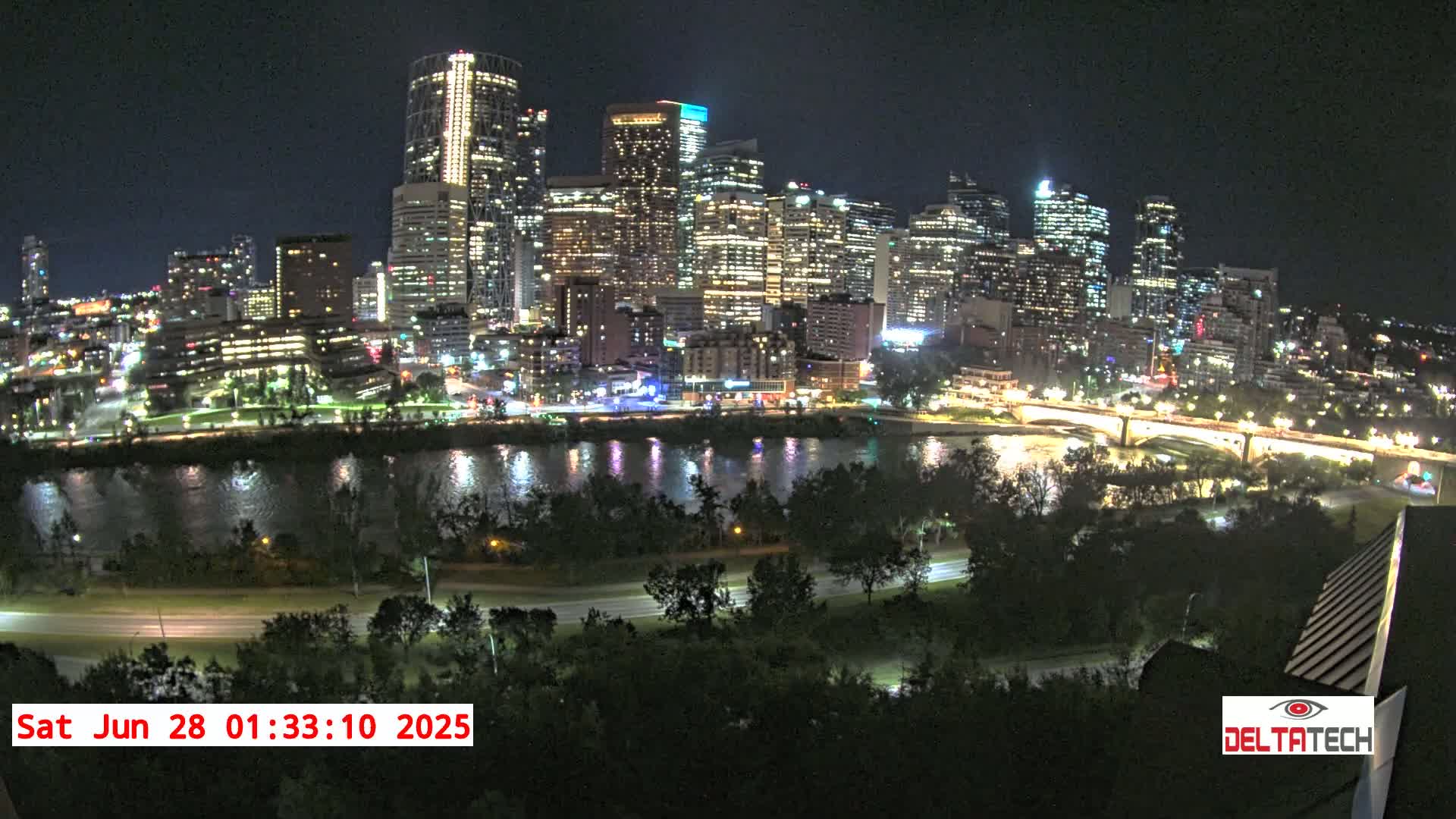 A brightly lit city skyline at night is seen across a dark river, with a road and trees in the foreground.