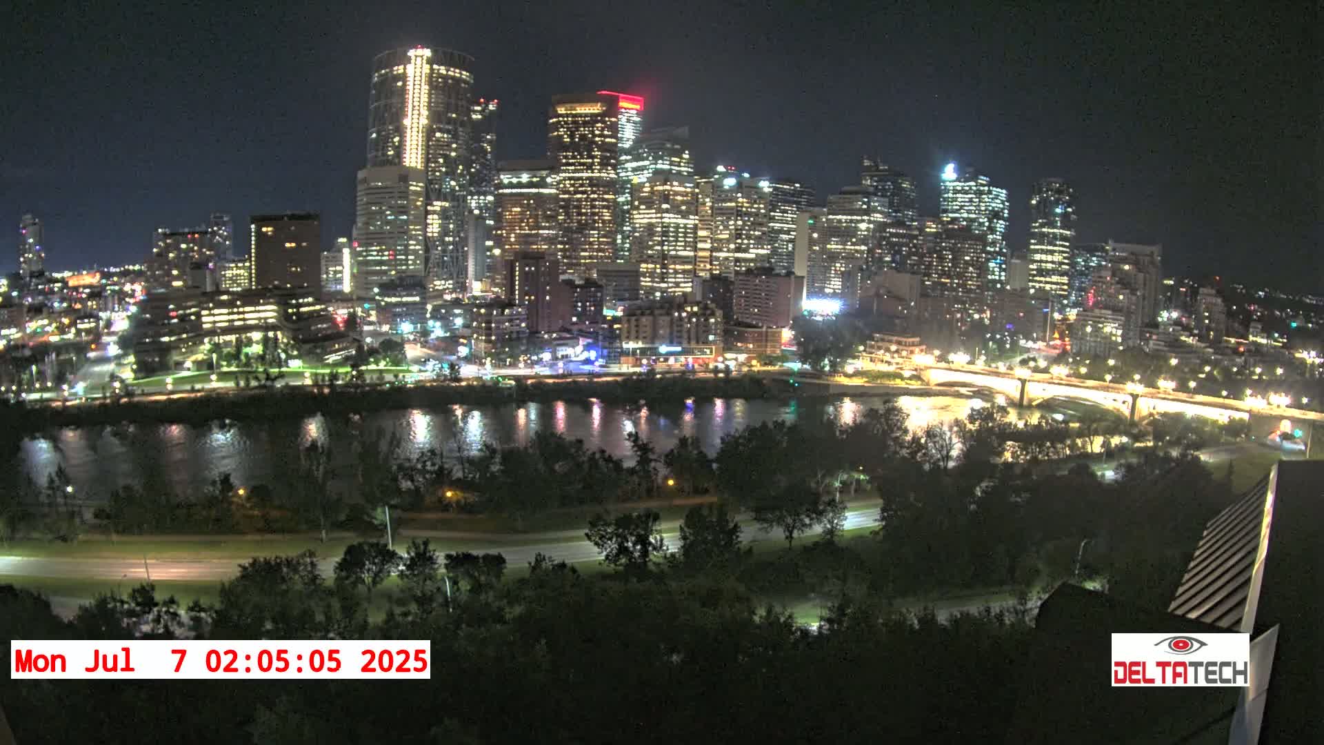 A brightly lit city skyline at night is seen across a calm river, bordered by trees and roads.