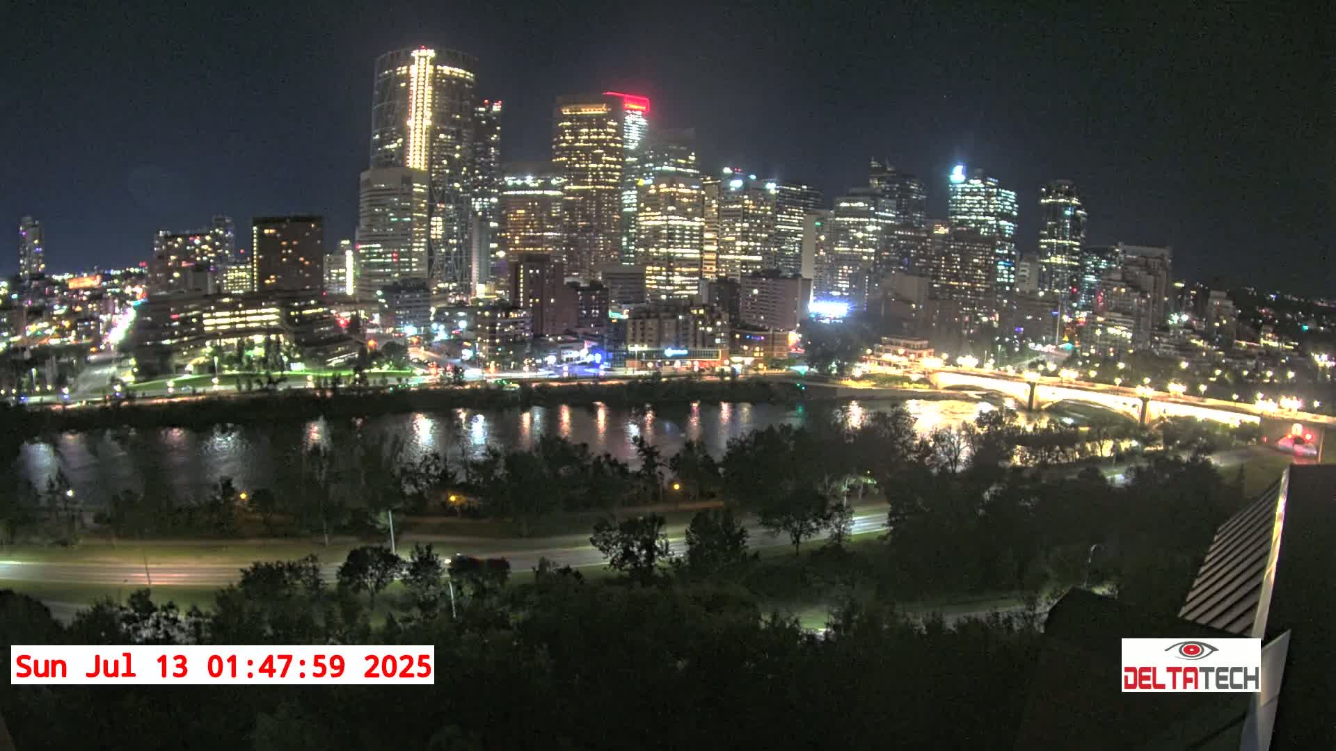 A brightly lit city skyline at night is seen across a calm river, with trees and a road in the foreground under a clear sky.