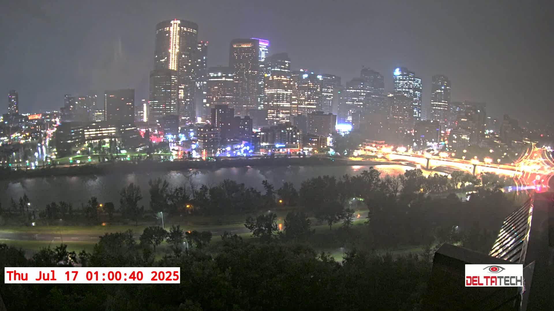 A nighttime view of a city skyline across a river, illuminated by lights, during a rain shower.
