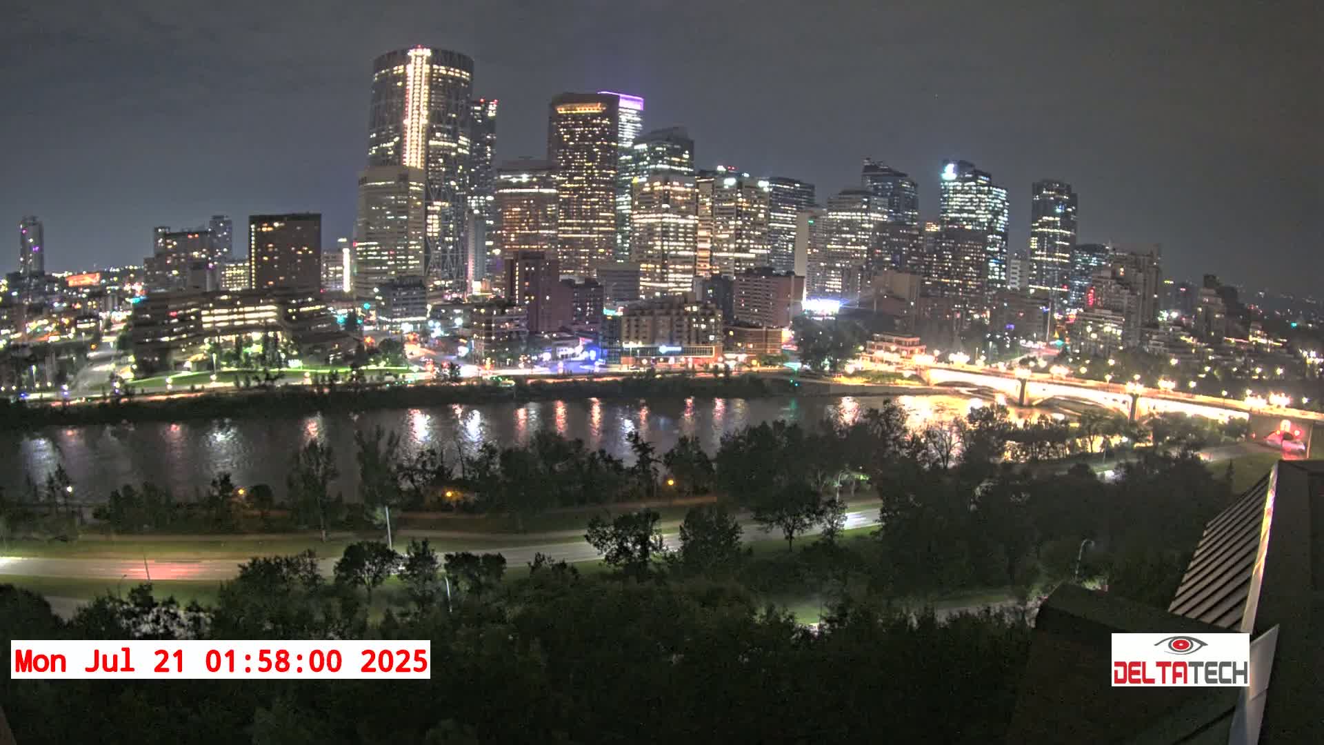 A nighttime view of a brightly lit city skyline across a dark river, with trees and a road in the foreground under a clear night sky.