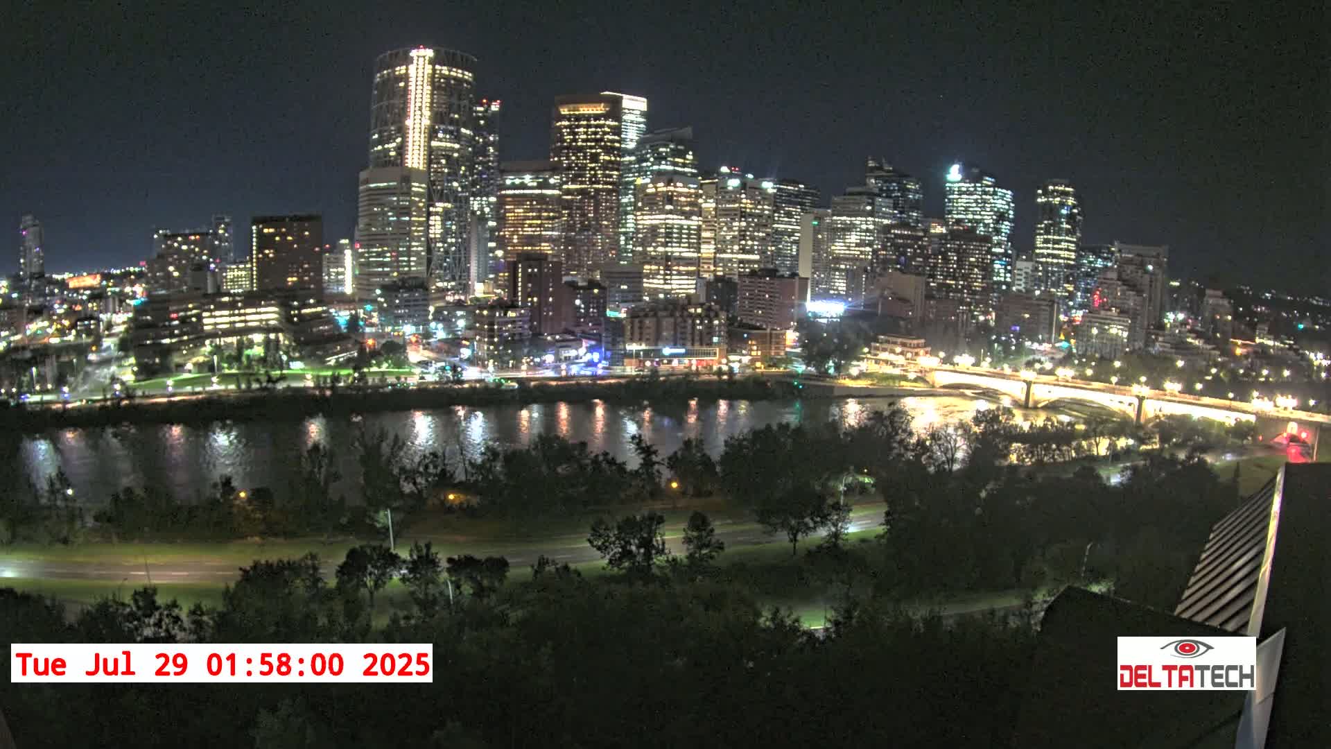 A brightly lit city skyline at night is seen across a dark river, with trees and a road in the foreground under a clear night sky.