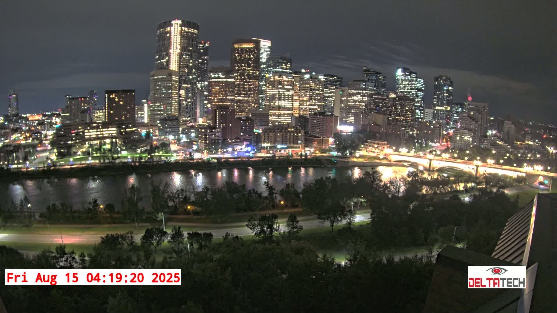 A brightly lit city skyline at night is seen across a dark river, with trees and a road in the foreground under a clear night sky.