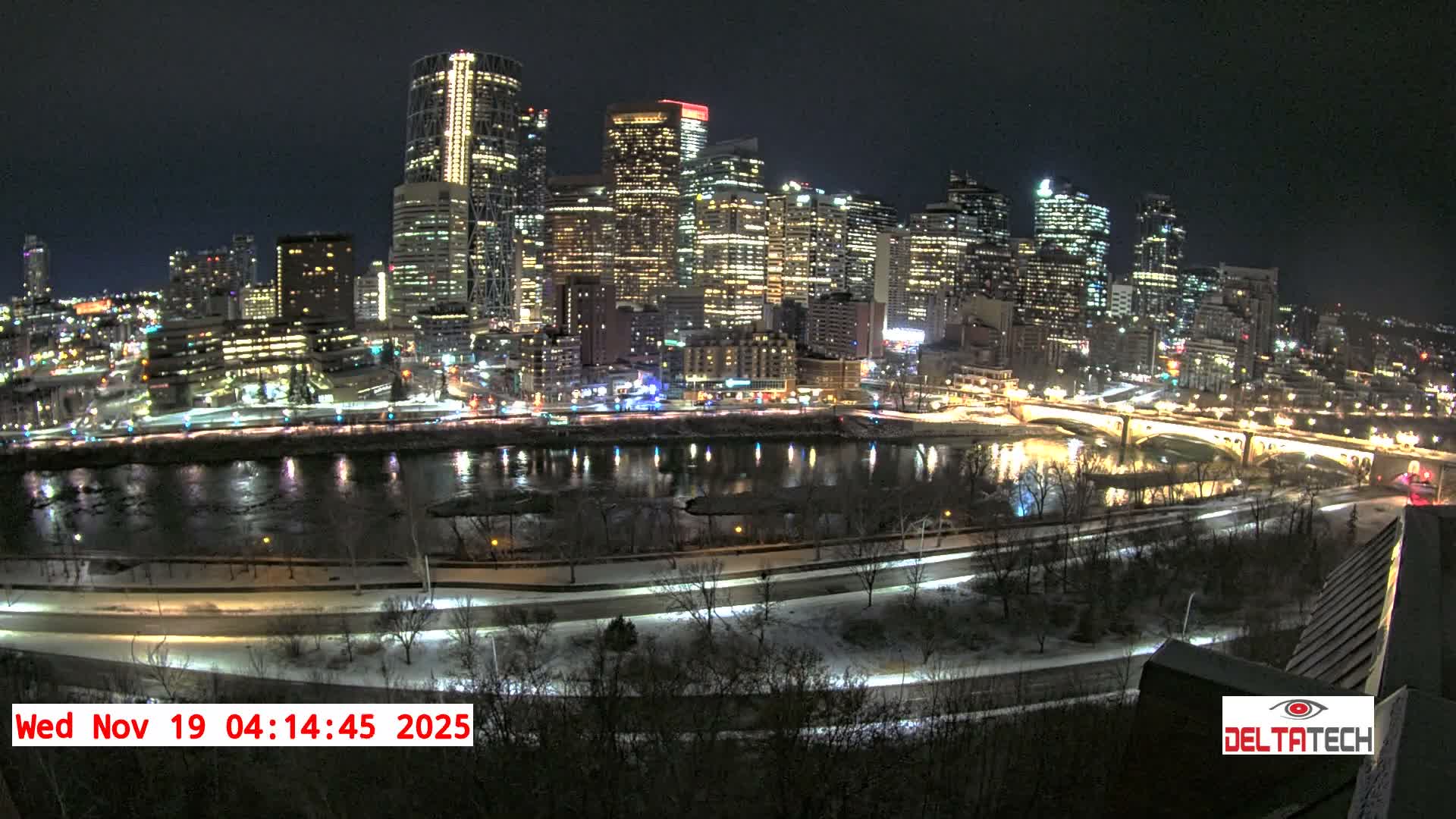 A brightly lit city skyline at night is seen across a dark river, with trees and a road in the foreground under a clear night sky.